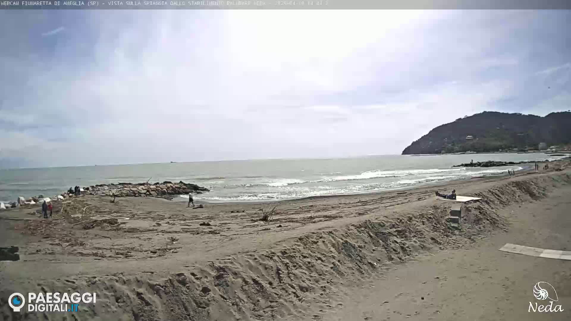 A sandy beach with a few people on it, a calm sea with small waves, and a hill in the background under a mostly cloudy sky.