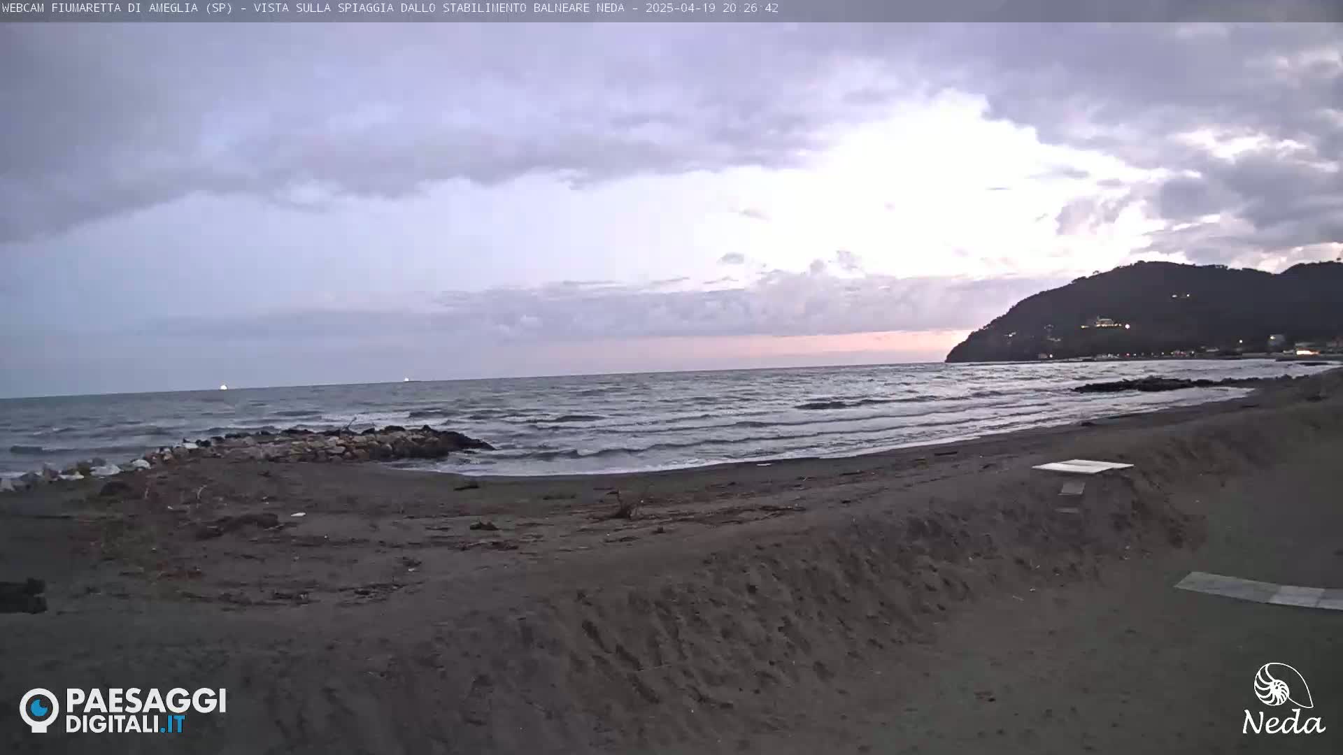 A dark, sandy beach with gentle waves under a cloudy, twilight sky, backed by a distant hill.