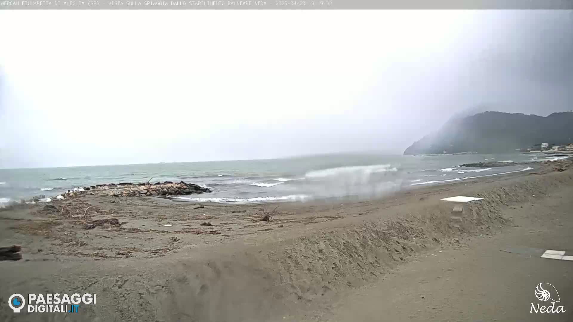 A sandy beach with moderate waves crashing on the shore under an overcast sky, with a distant mountain visible.