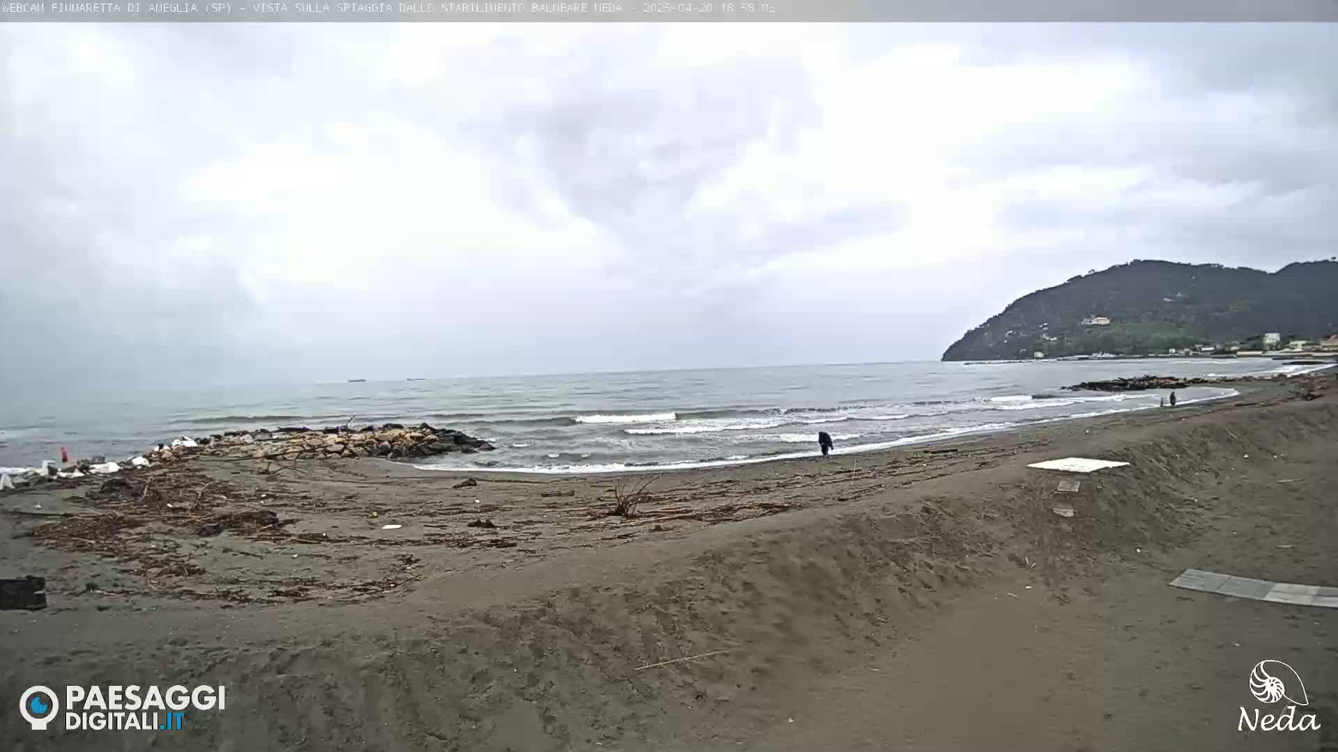 A dark-sand beach with small waves and a person walking on it, under an overcast sky, is backed by a hill.