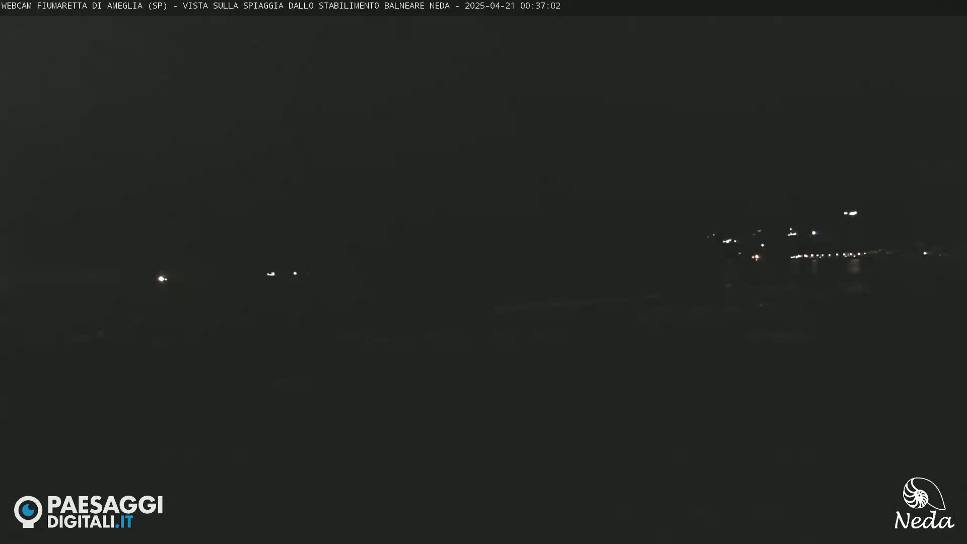 A dark nighttime view of a beach with distant lights visible across the water.