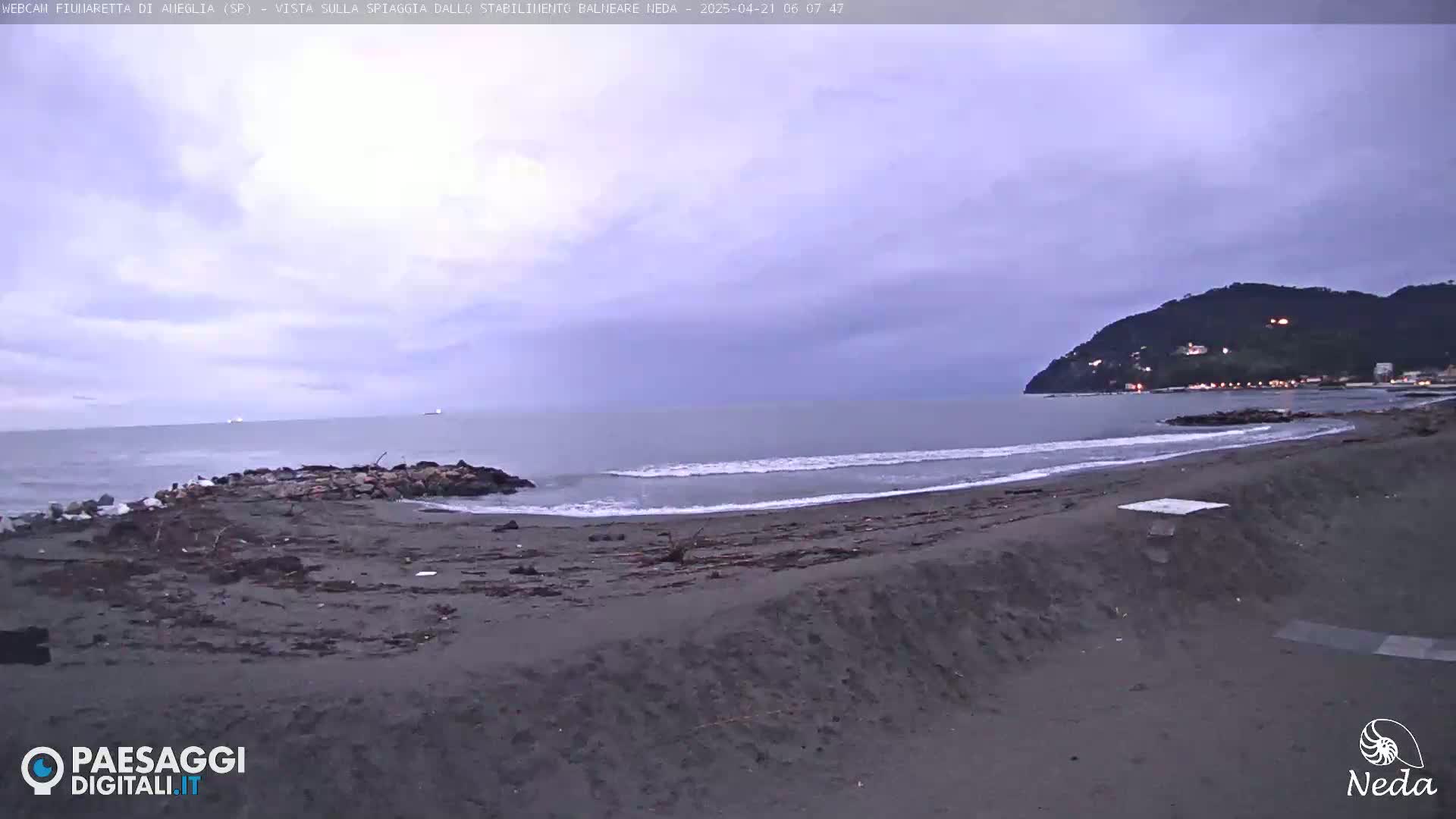 A dark, sandy beach with gentle waves under an overcast sky, a distant hill with lights visible on the coast.