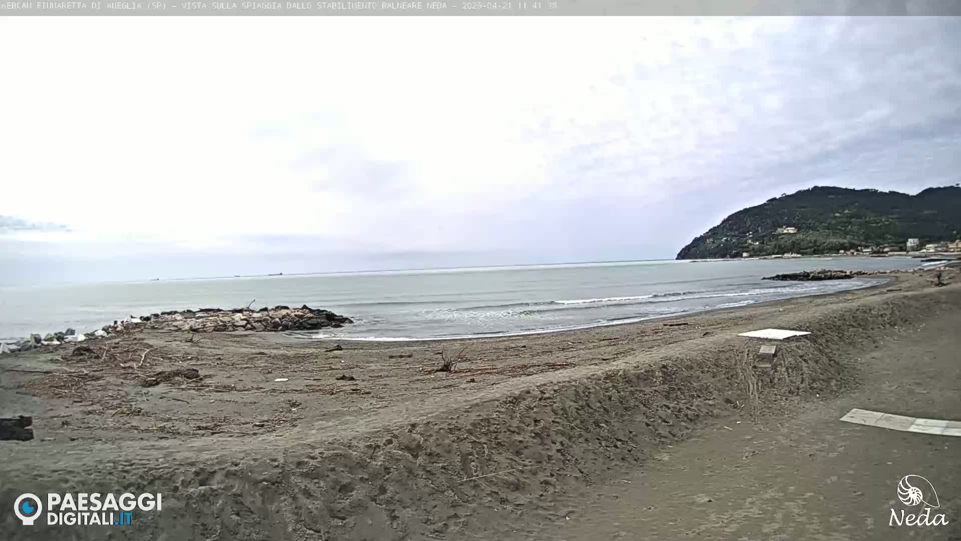 A mostly cloudy day shows a wide sandy beach with a calm sea and a distant hill in the background.