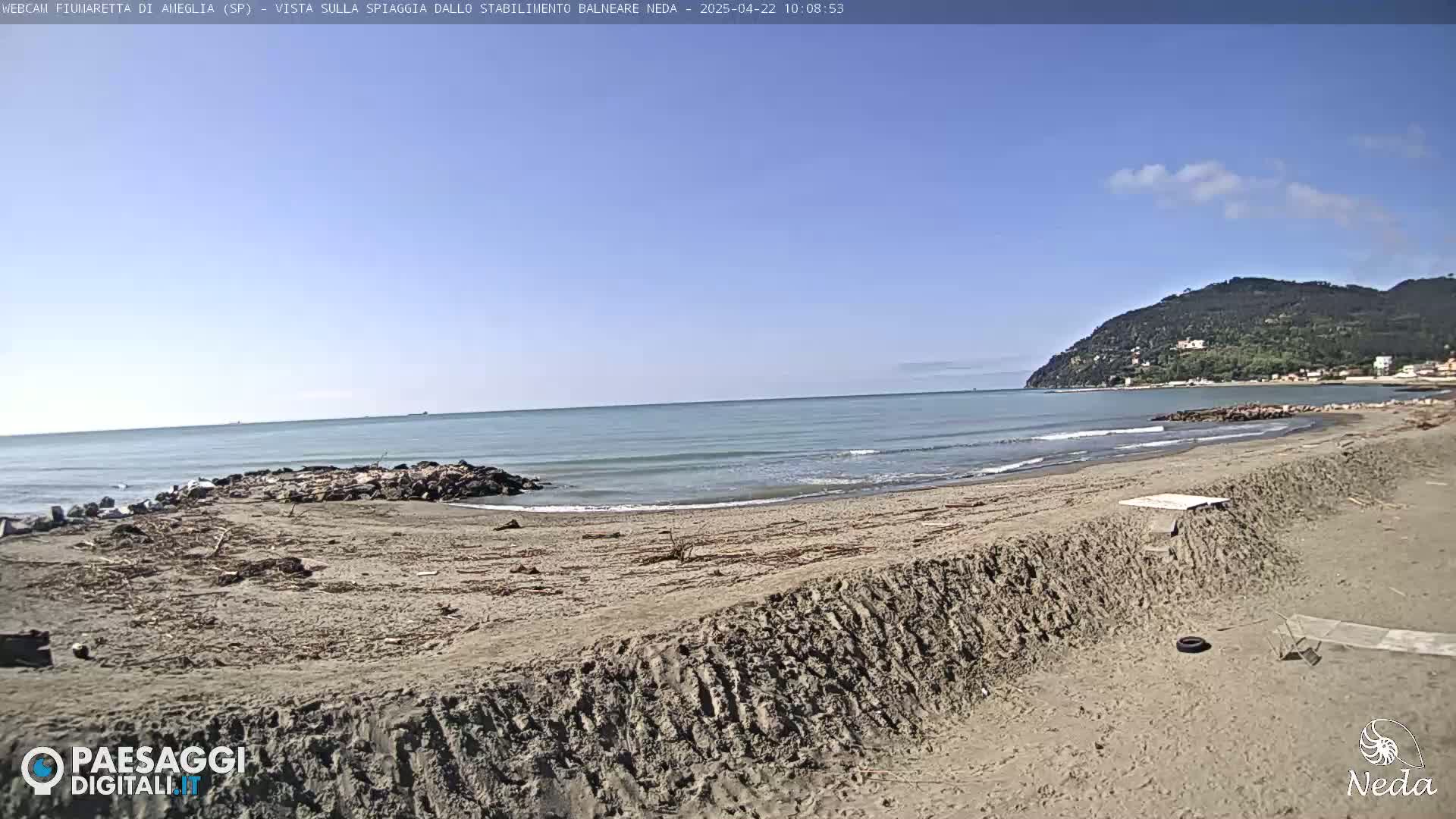 A sandy beach with a calm sea under a clear blue sky shows a rocky outcrop near the water's edge and a hill in the distance.