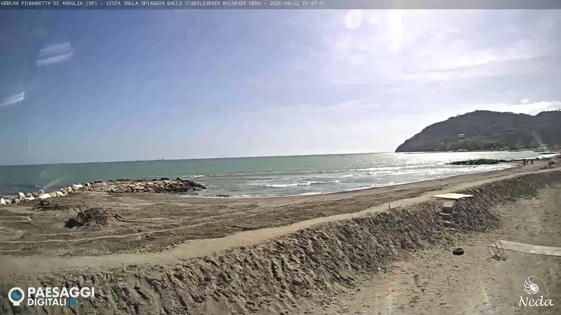A sandy beach with a calm sea under a mostly sunny sky, backed by a low hill.