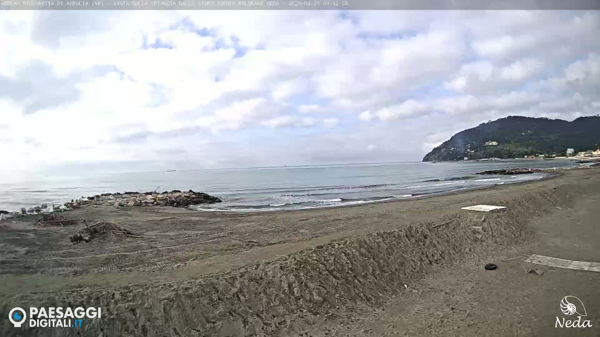 A partly cloudy day reveals a sandy beach with a calm sea and a distant hill under a mostly cloudy sky.