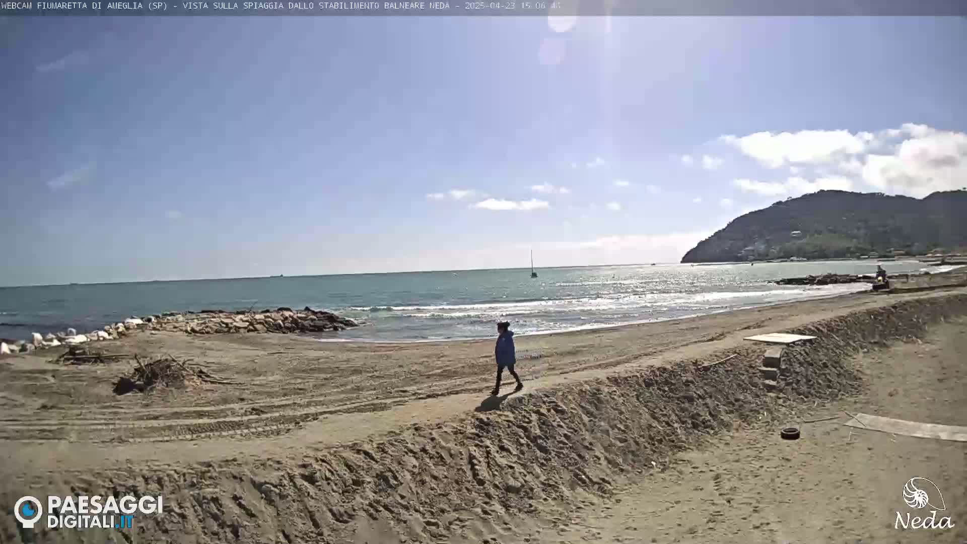 A person walks along a sandy beach by a calm sea under a mostly sunny sky, with a hill visible in the distance.