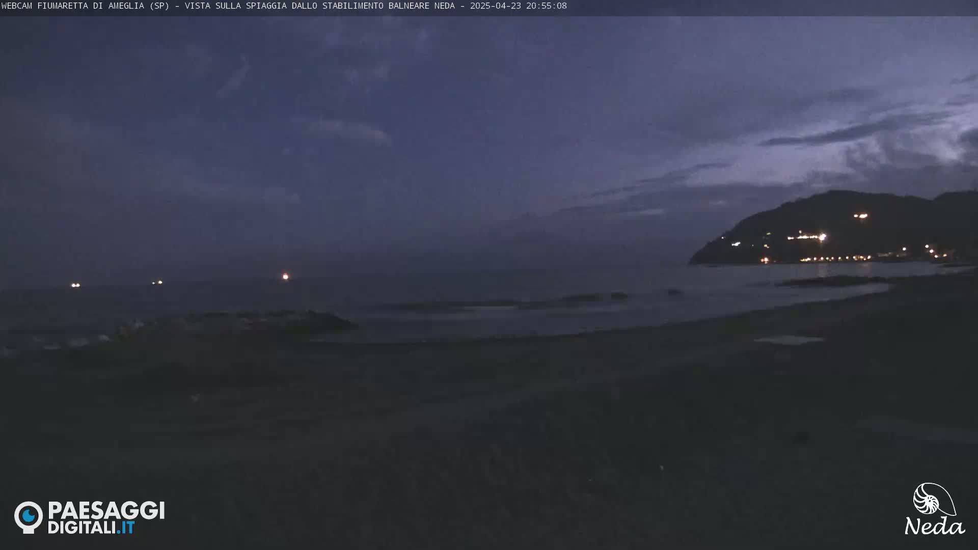A dark, twilight view of a beach at night, with a hill in the distance illuminated by lights and a few distant lights on the water.
