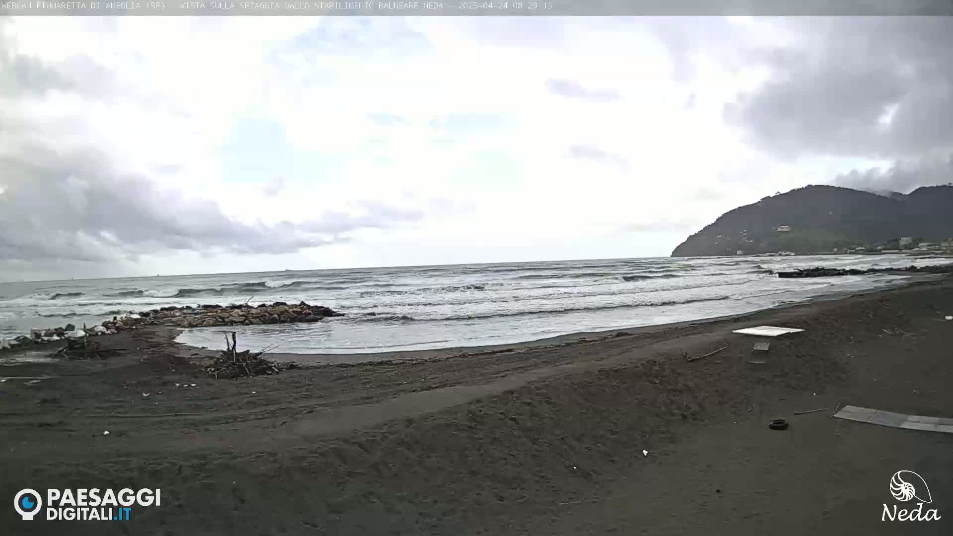 A dark-sand beach with moderate waves under an overcast sky, backed by a dark, tree-covered hill.