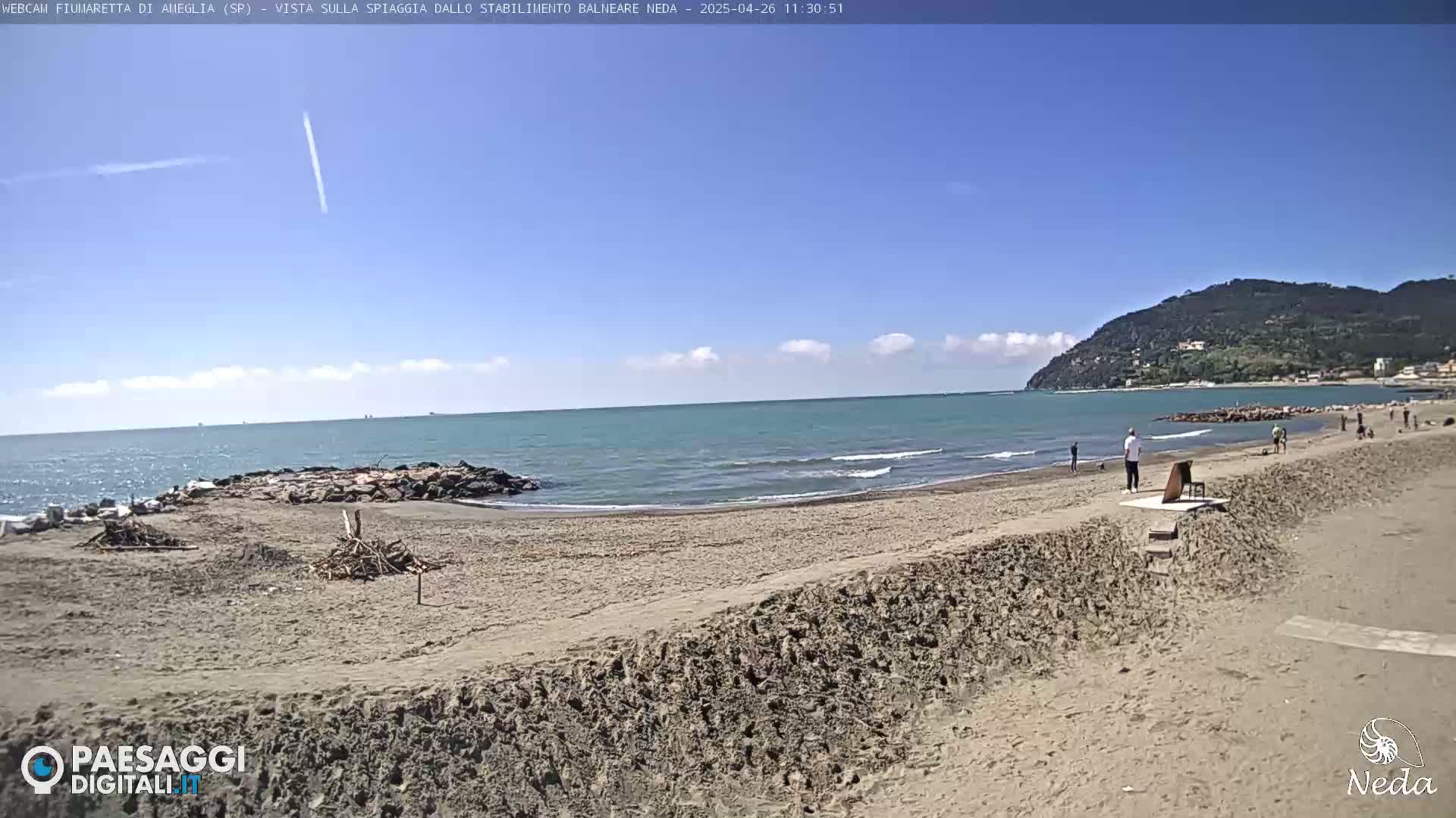 A mostly sunny day at a sandy beach shows a calm sea with some small waves, a few people on the shore, and a distant hill.