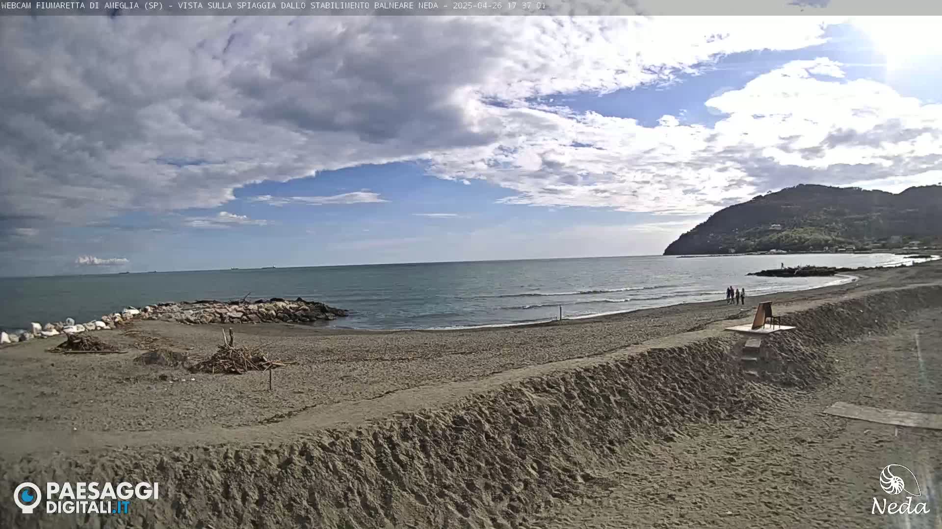 A mostly cloudy day at a sandy beach with calm ocean waves, a rocky jetty, and a hill in the background.