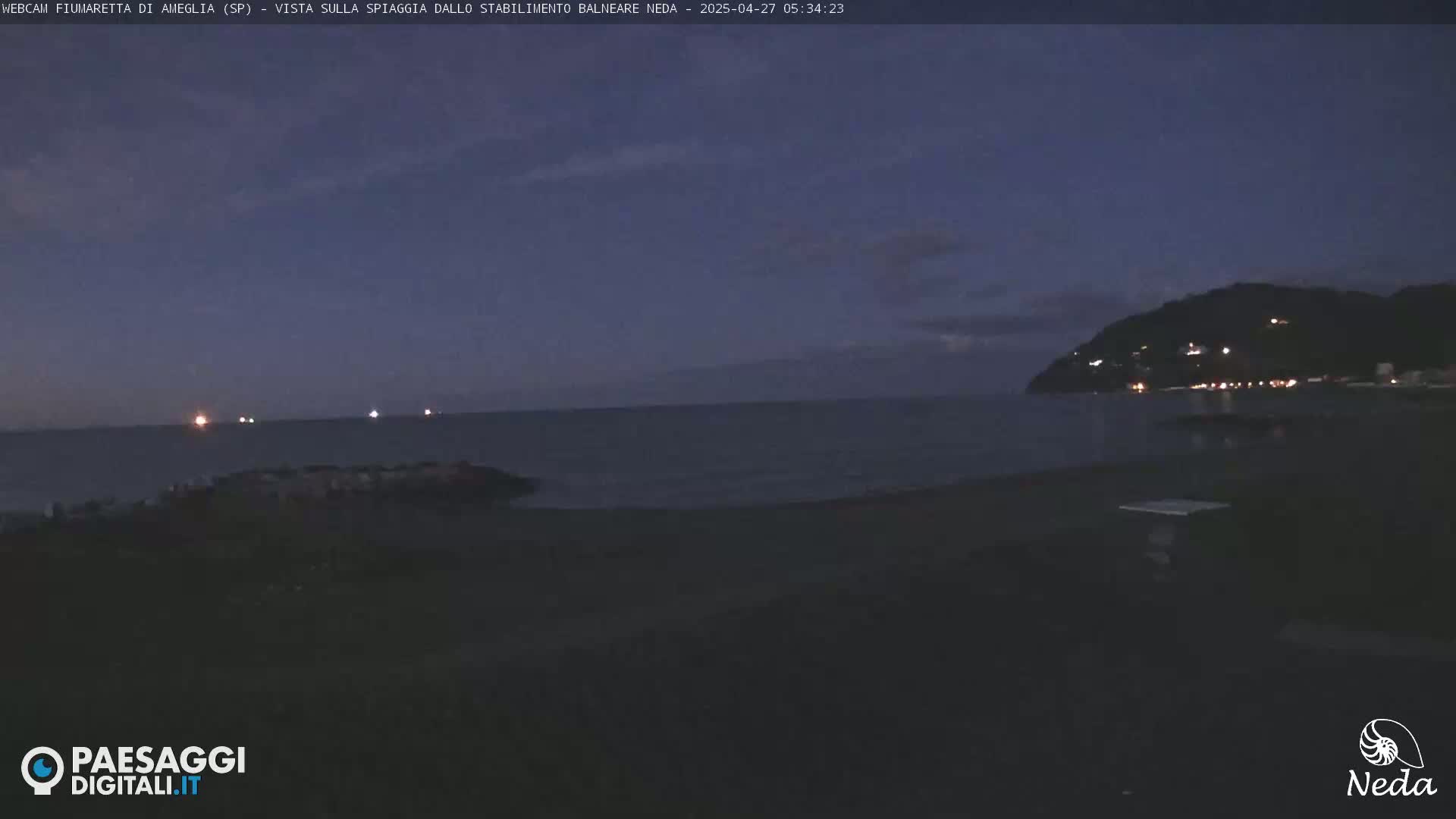 A dark, twilight view of a beach and calm ocean, with a hill in the background showing some lights.