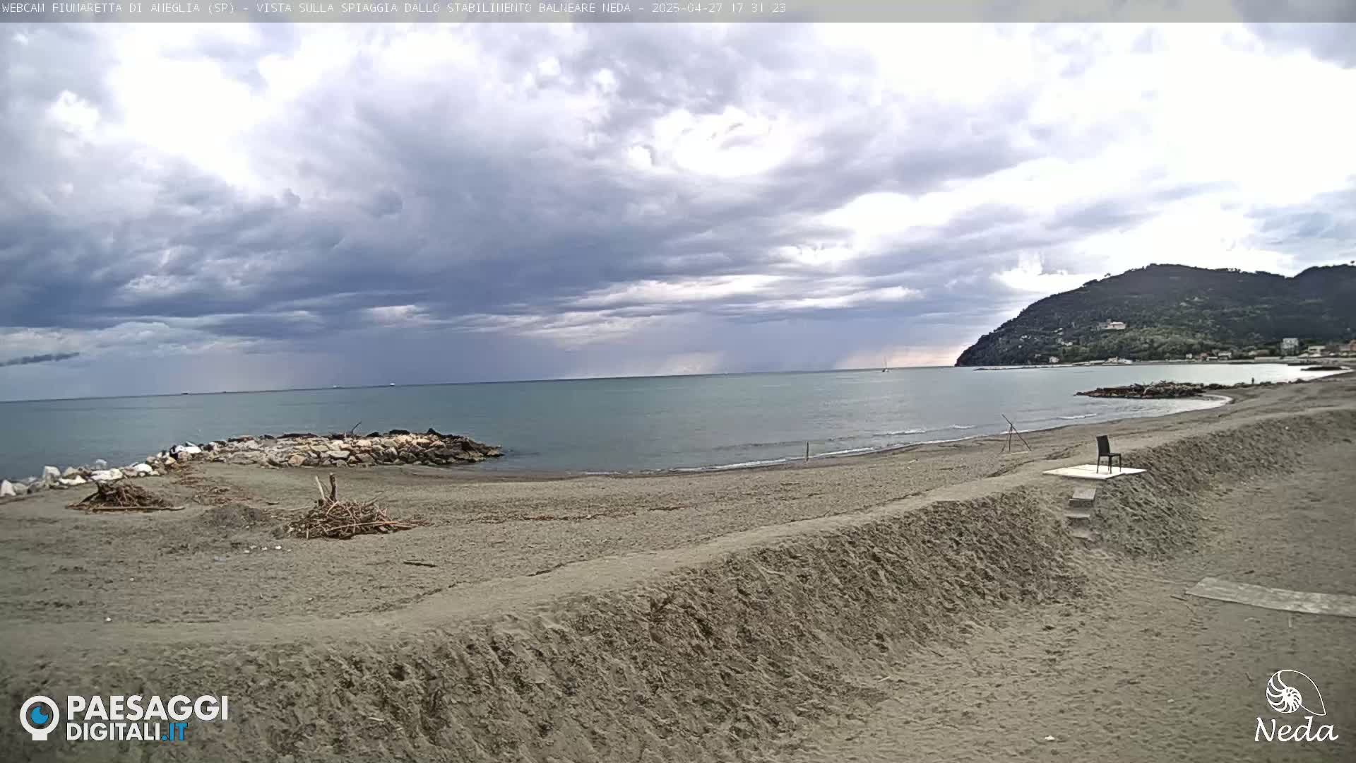 A sandy beach with a single chair sits on a small platform with steps leading to it, under a cloudy sky, overlooking calm water and a distant hill.