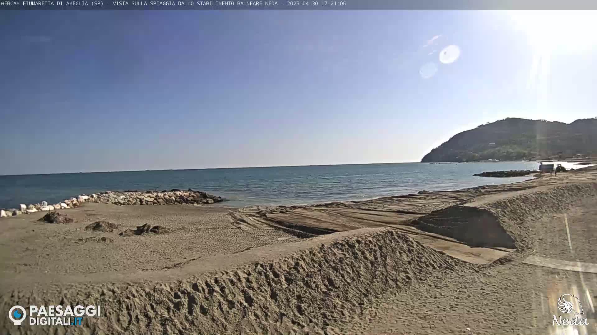 A sandy beach with tire tracks and a rocky breakwater extends to a calm sea under a clear, sunny sky, backed by a distant hill.