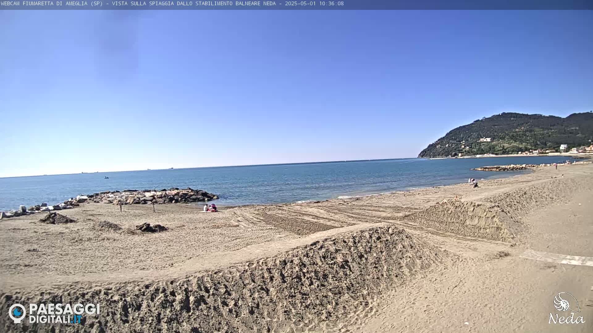 A sandy beach with a calm ocean under a clear blue sky, and a hill in the background.