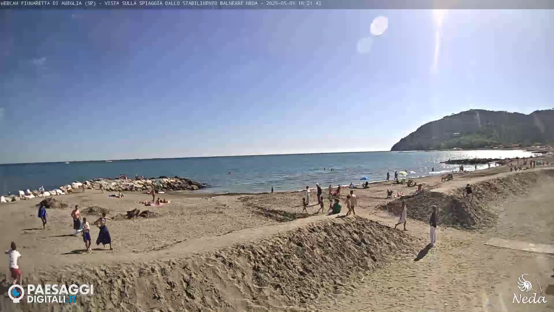 A sunny day at a sandy beach shows people swimming and relaxing near a calm sea with a hill in the background.