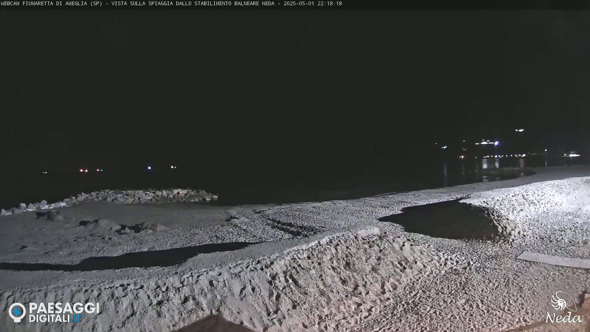 A nighttime view of a sandy beach at the edge of a dark body of water, with lights visible in the distance.