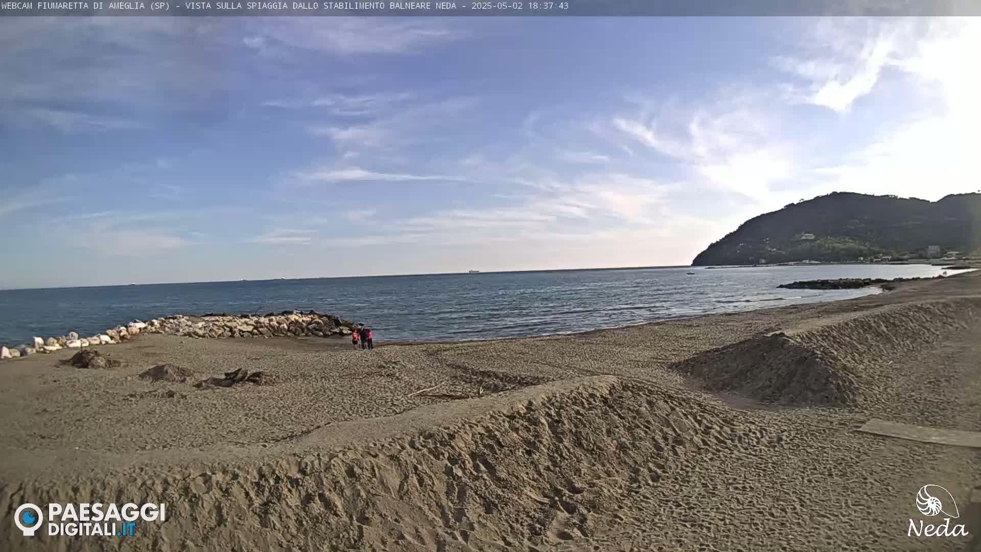 A sandy beach stretches to a calm sea under a partly cloudy sky, with a distant hill visible on the right.