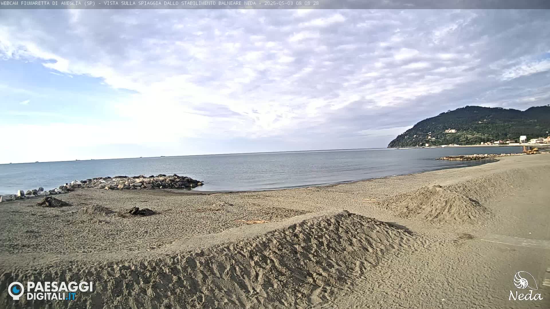 A mostly cloudy day reveals a sandy beach with piles of sand, a calm sea, and a distant, tree-covered hill.