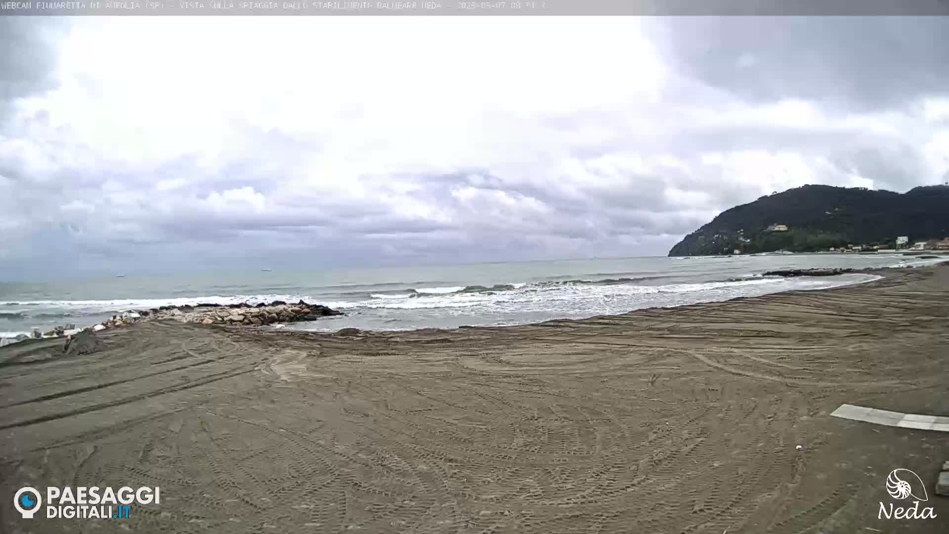 A mostly cloudy day at a sandy beach with small waves lapping the shore, and a distant hill visible in the background.