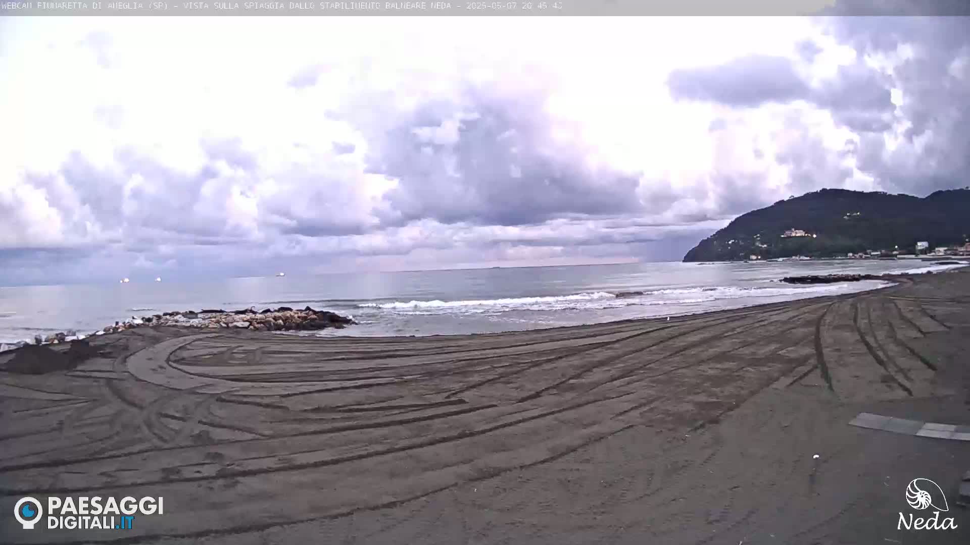 A dark-sand beach with tire tracks is visible, extending to a somewhat choppy sea under a cloudy sky; a distant hill is visible on the right.
