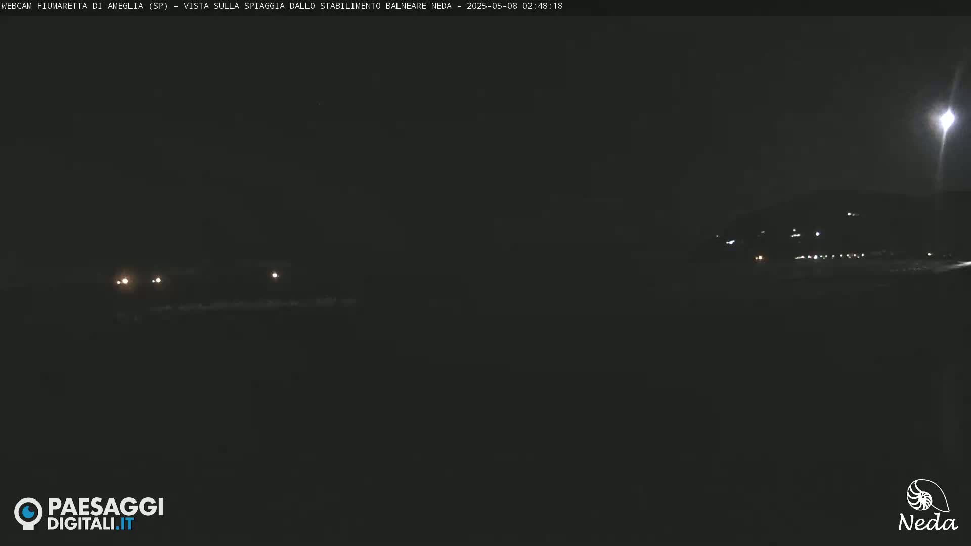A dark nighttime scene shows a few distant lights on a beach and hilltop, under a mostly clear sky.