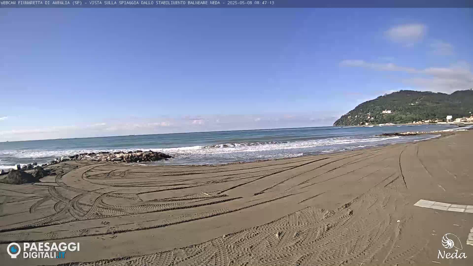 A sandy beach with tire tracks shows gentle waves lapping the shore under a clear blue sky.