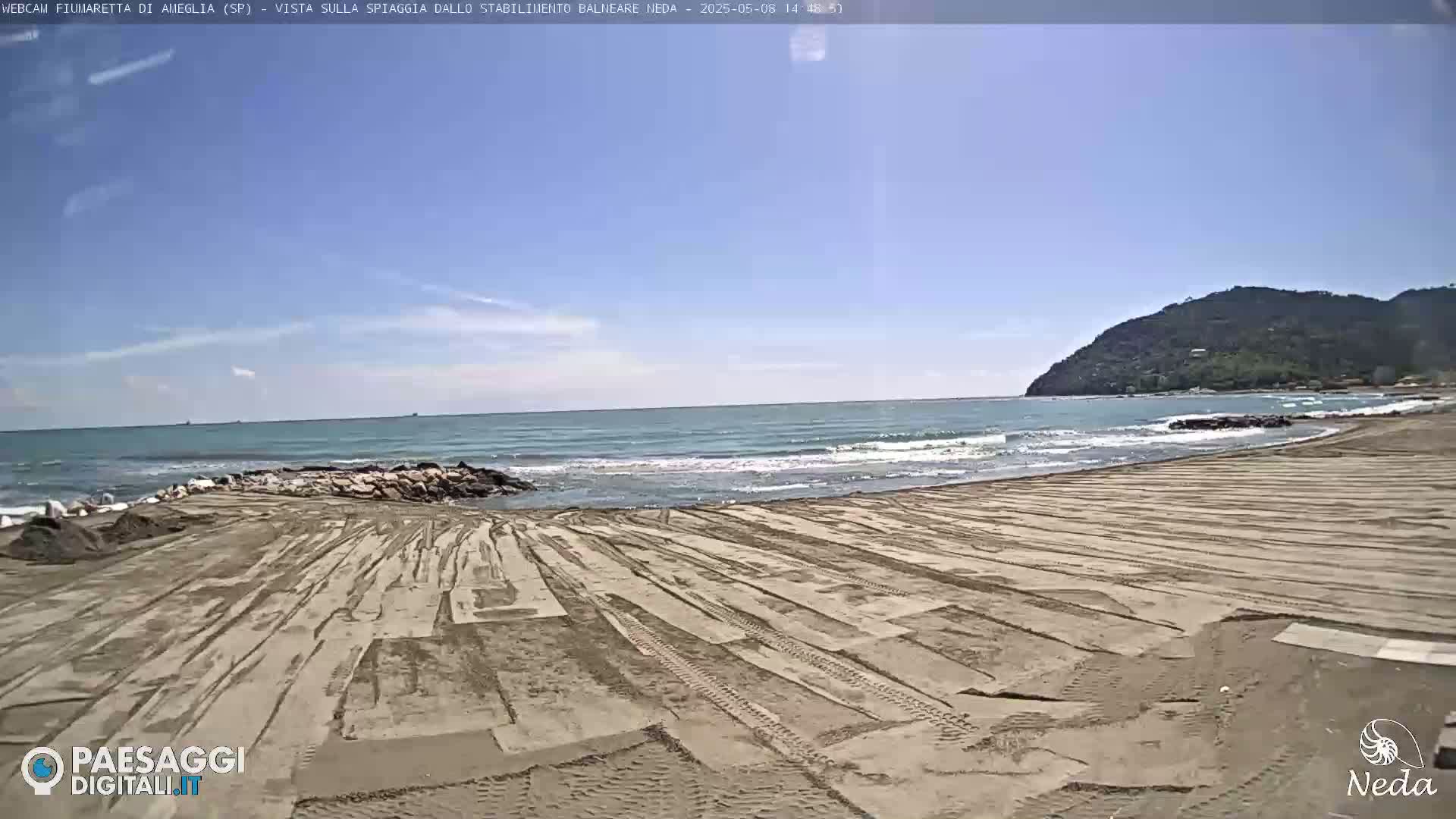 A sandy beach with tire tracks stretches toward a calm sea under a clear blue sky, with a distant hill in the background.