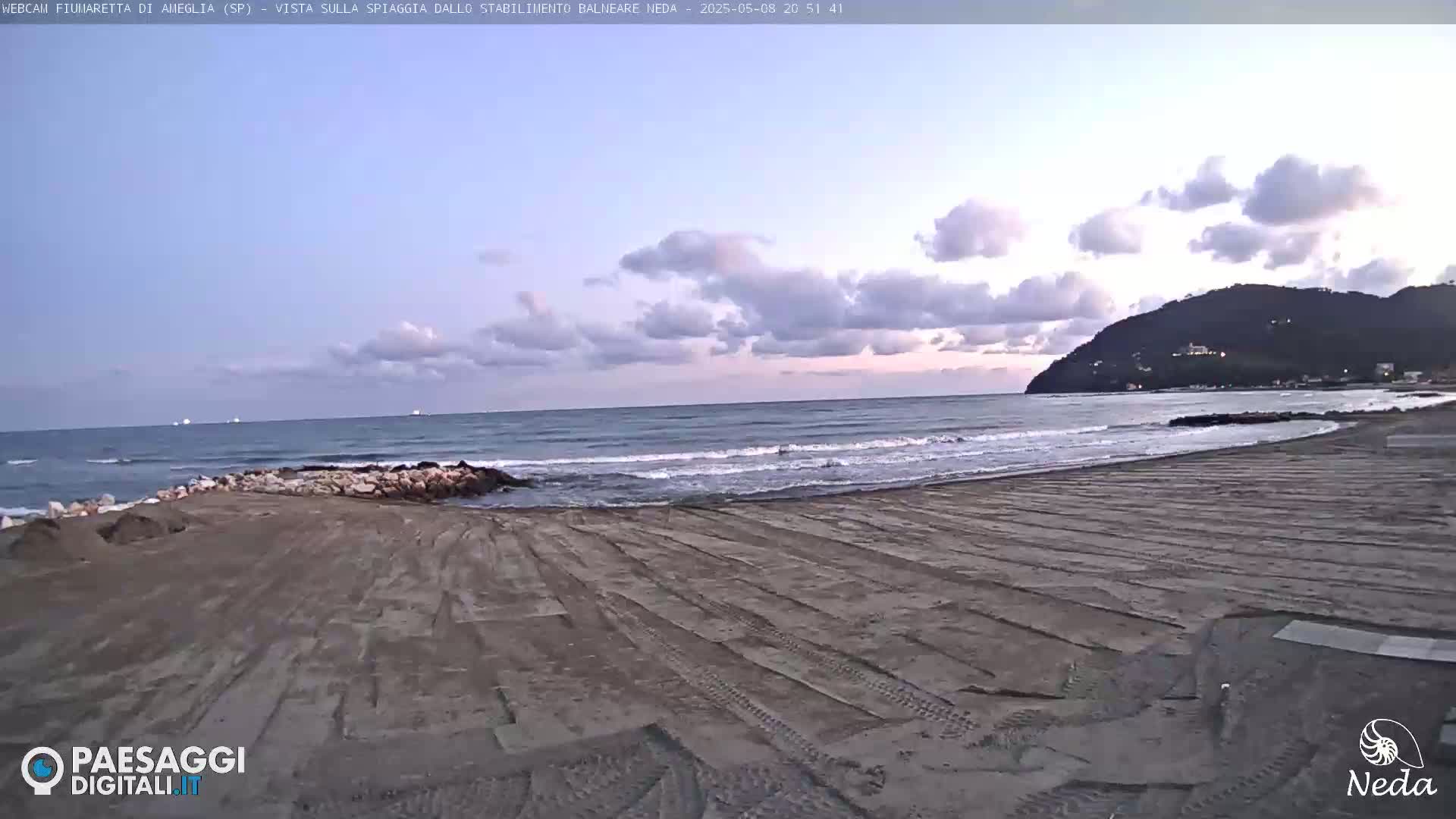 A sandy beach with tire tracks stretches toward a calm sea under a partly cloudy twilight sky, backed by a distant hill.