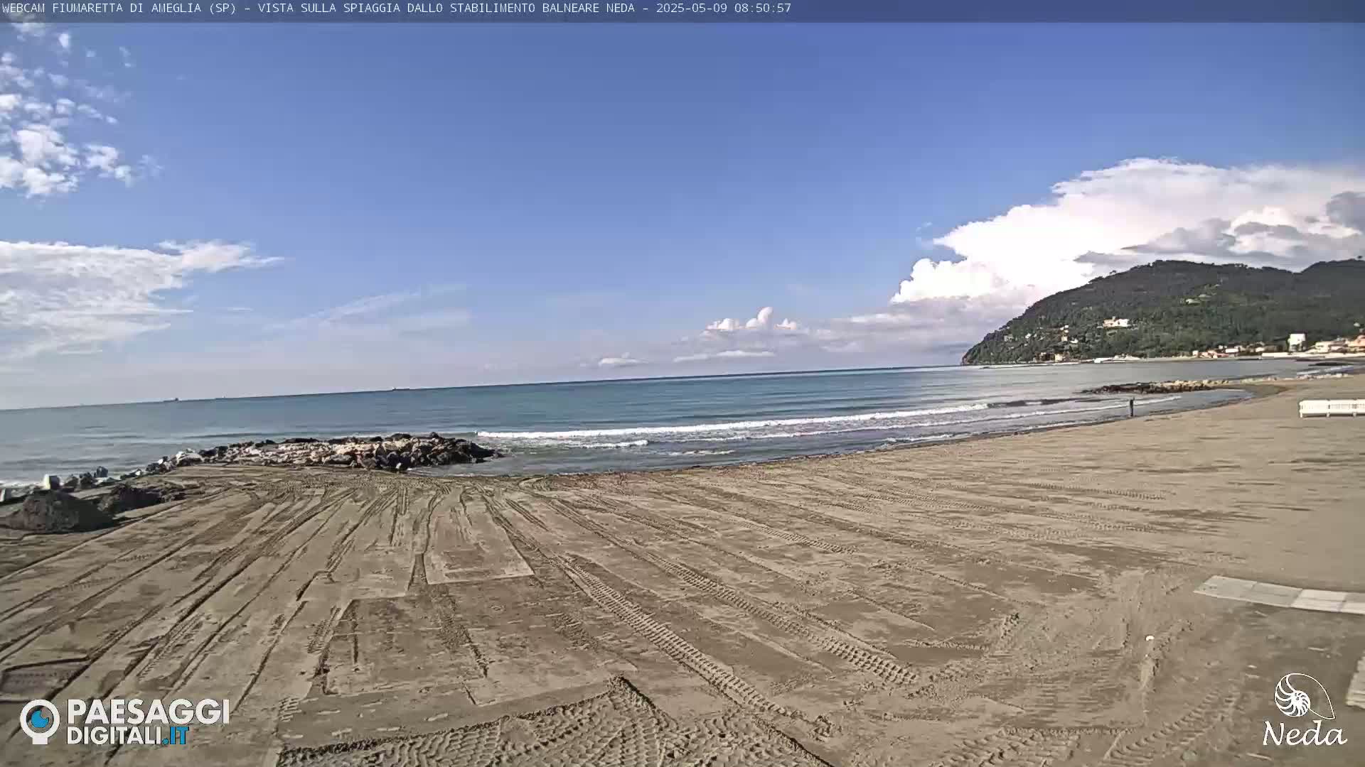 A sandy beach with tire tracks, a calm sea with small waves, and a partly cloudy sky, features a rocky area near the water and a hill in the distance.