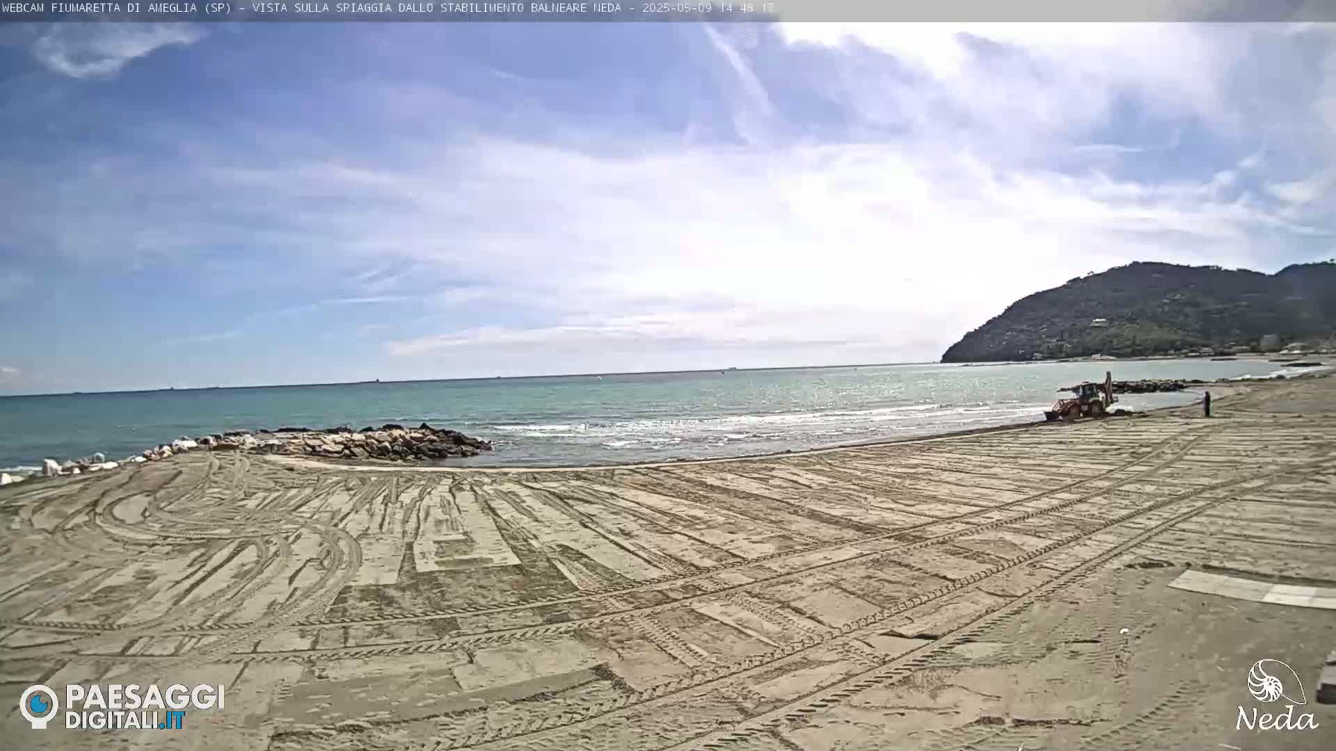 A sandy beach with tire tracks is being groomed by a piece of machinery under a partly cloudy sky, with a calm sea in the background and a hill visible in the distance.