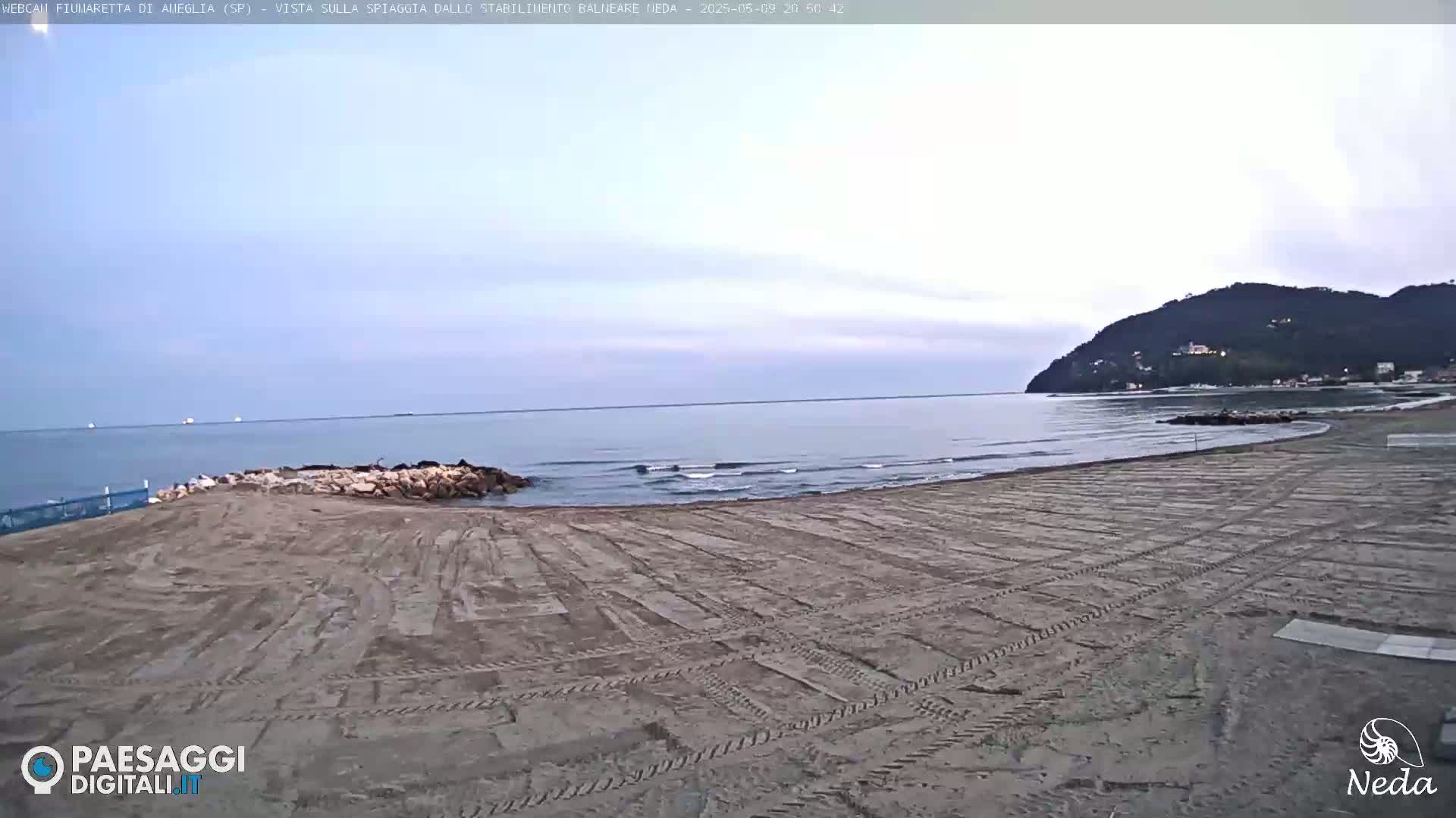 A mostly empty sandy beach with tire tracks stretches towards a calm sea under a pale, overcast sky, backed by a distant hill.
