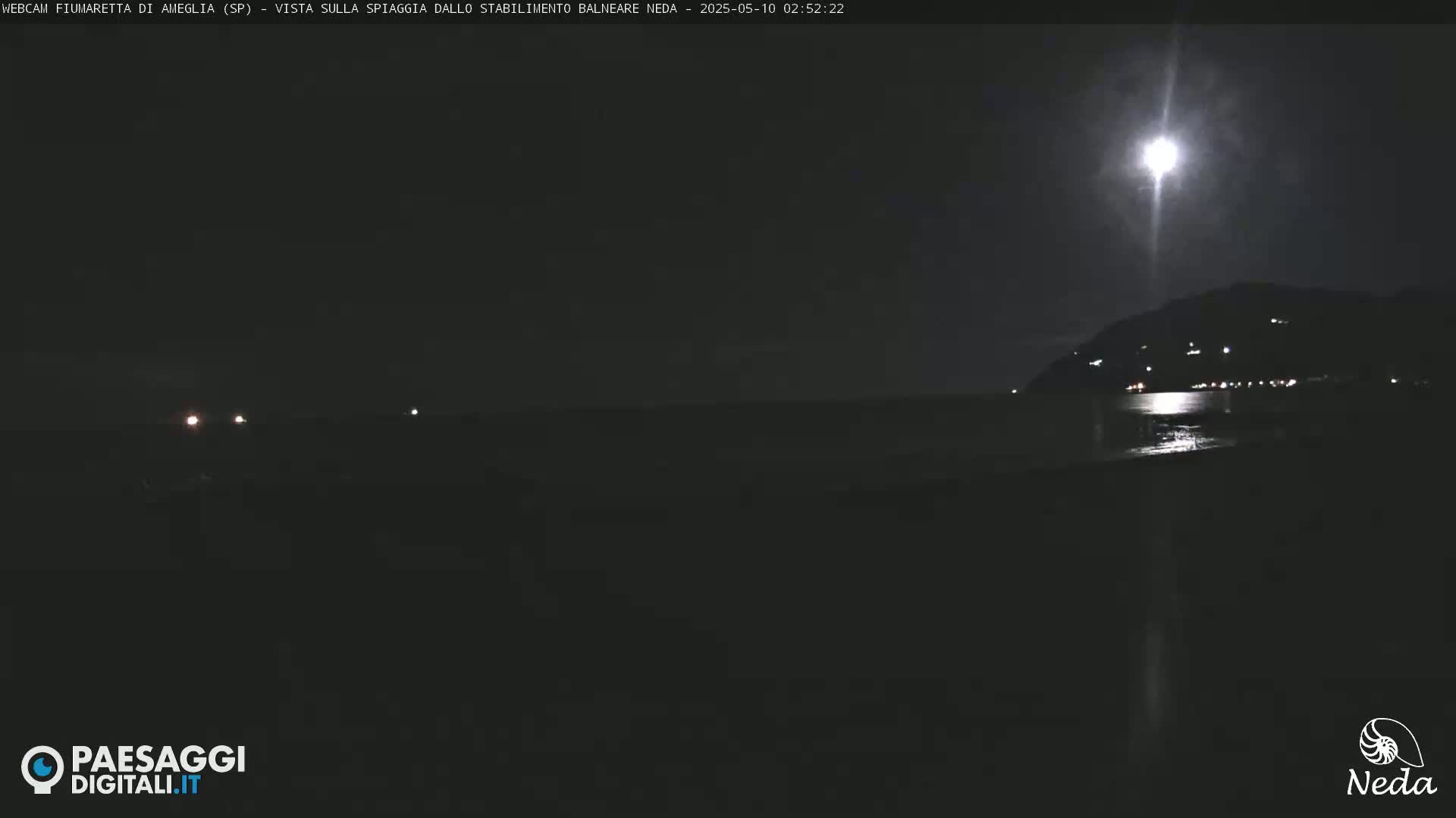A bright moon illuminates a dark beach and distant, dimly lit hillside at night.