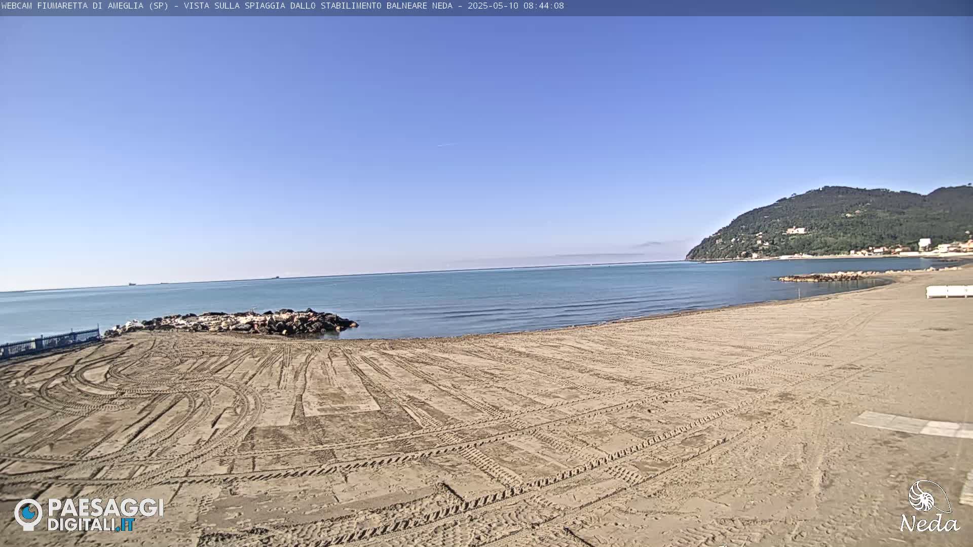 A sandy beach with tire tracks stretches towards a calm, blue sea under a clear sky, backed by a distant, green-covered hill.