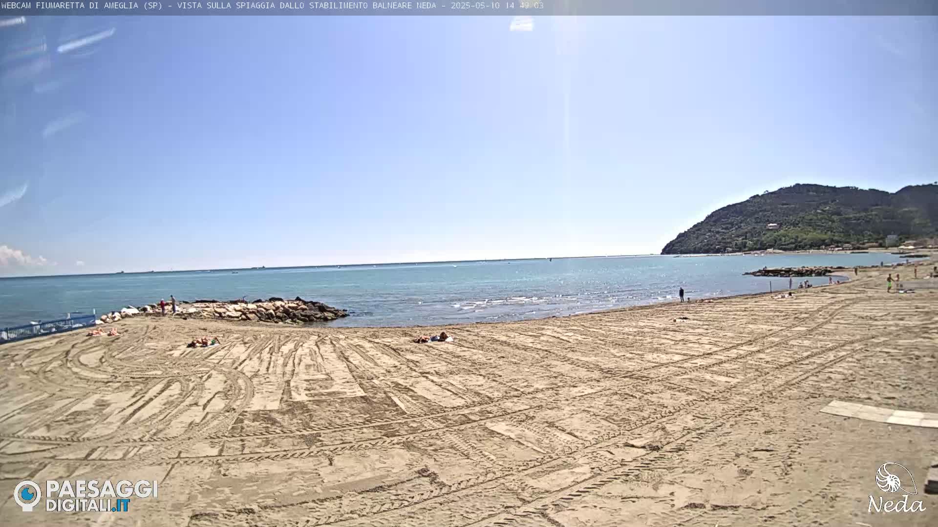 A sandy beach with a few people on it stretches out to a calm sea under a clear, sunny sky, backed by a low, green-covered hill.
