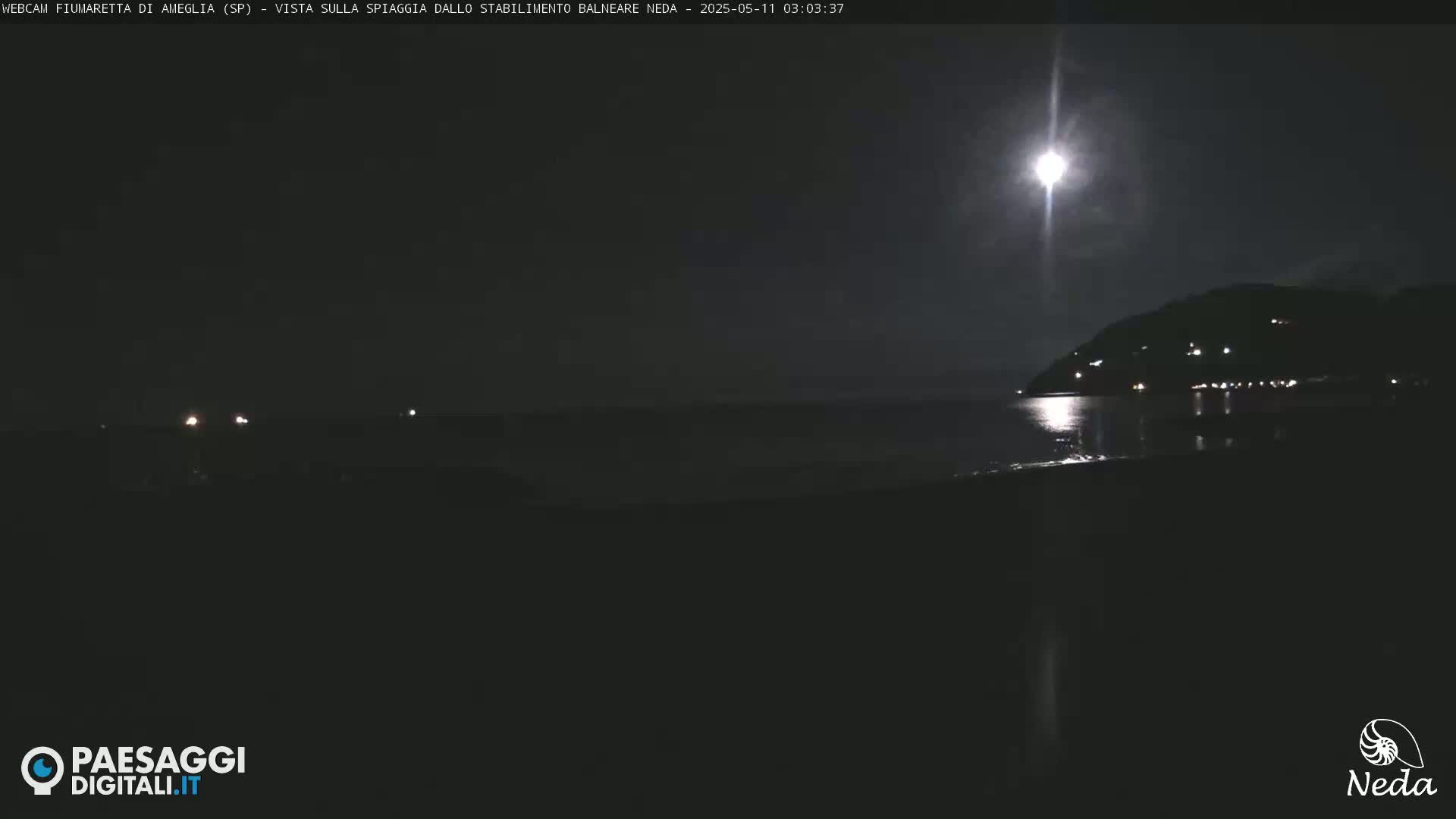 A bright moon illuminates a dark coastline and calm sea at night.