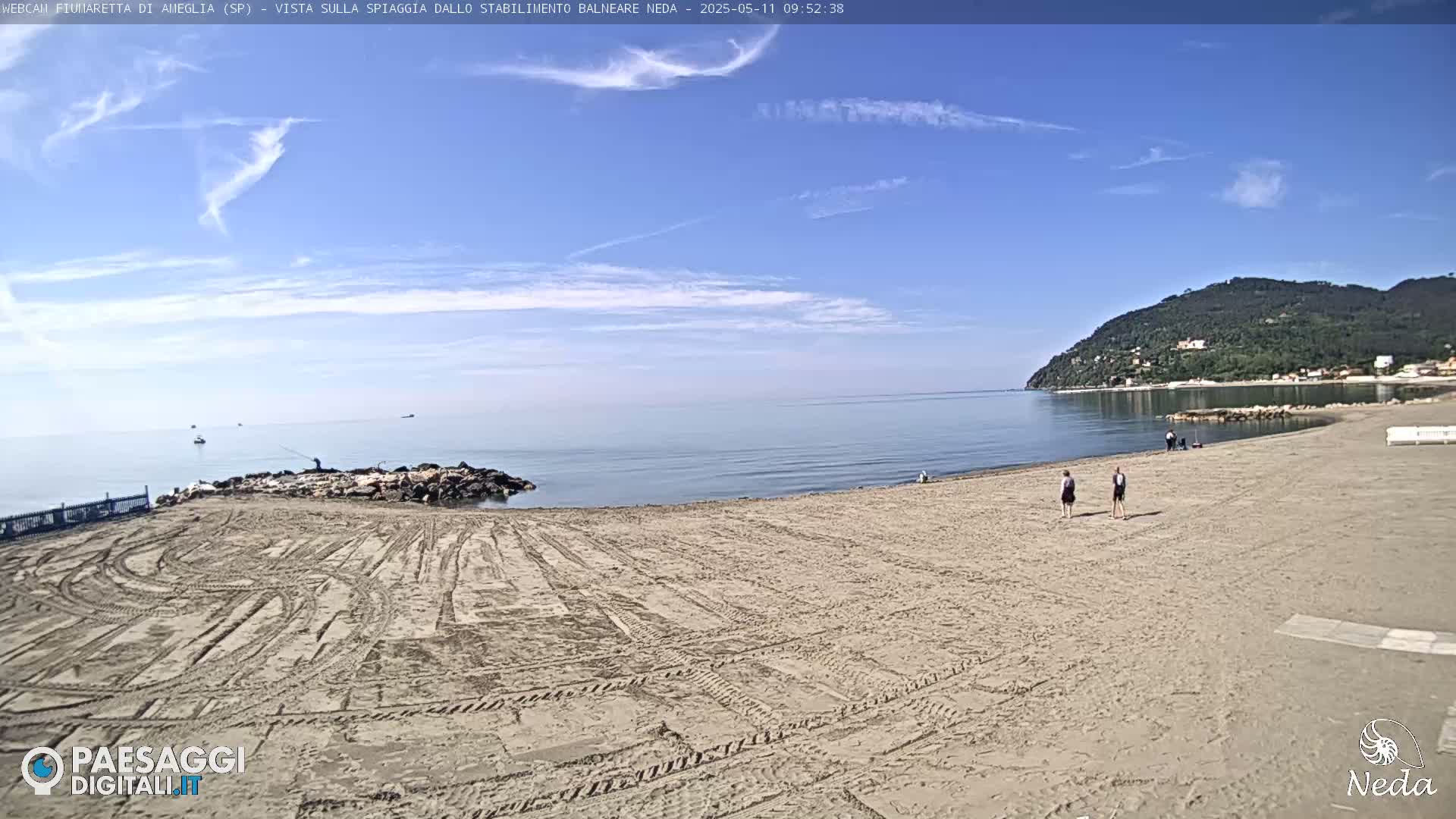A sandy beach stretches toward a calm sea under a mostly clear blue sky with a few wispy clouds, and a green-covered hill is visible in the distance.