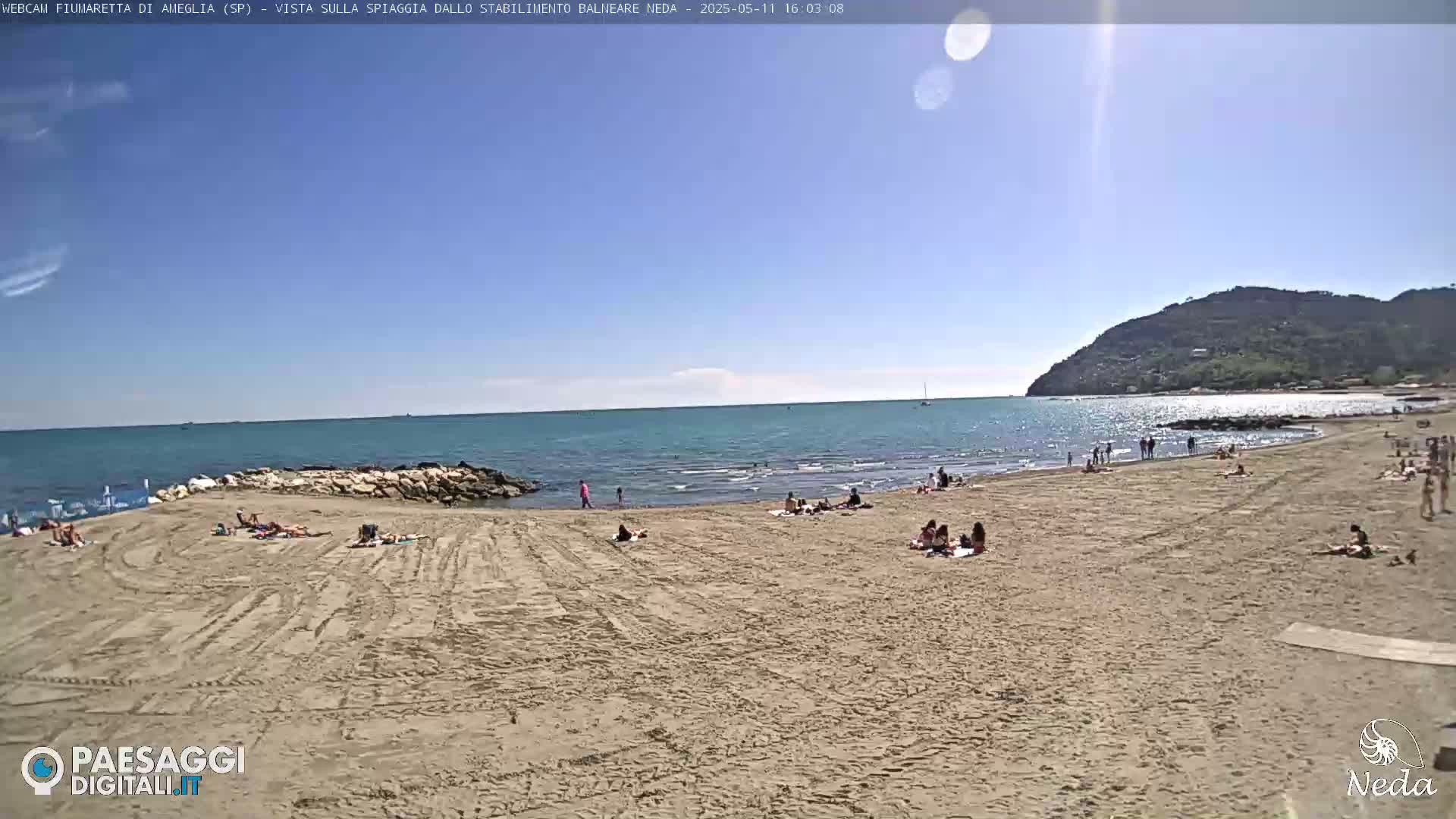 A sandy beach with scattered sunbathers and calm, turquoise water under a clear, sunny sky, backed by a rocky hill.