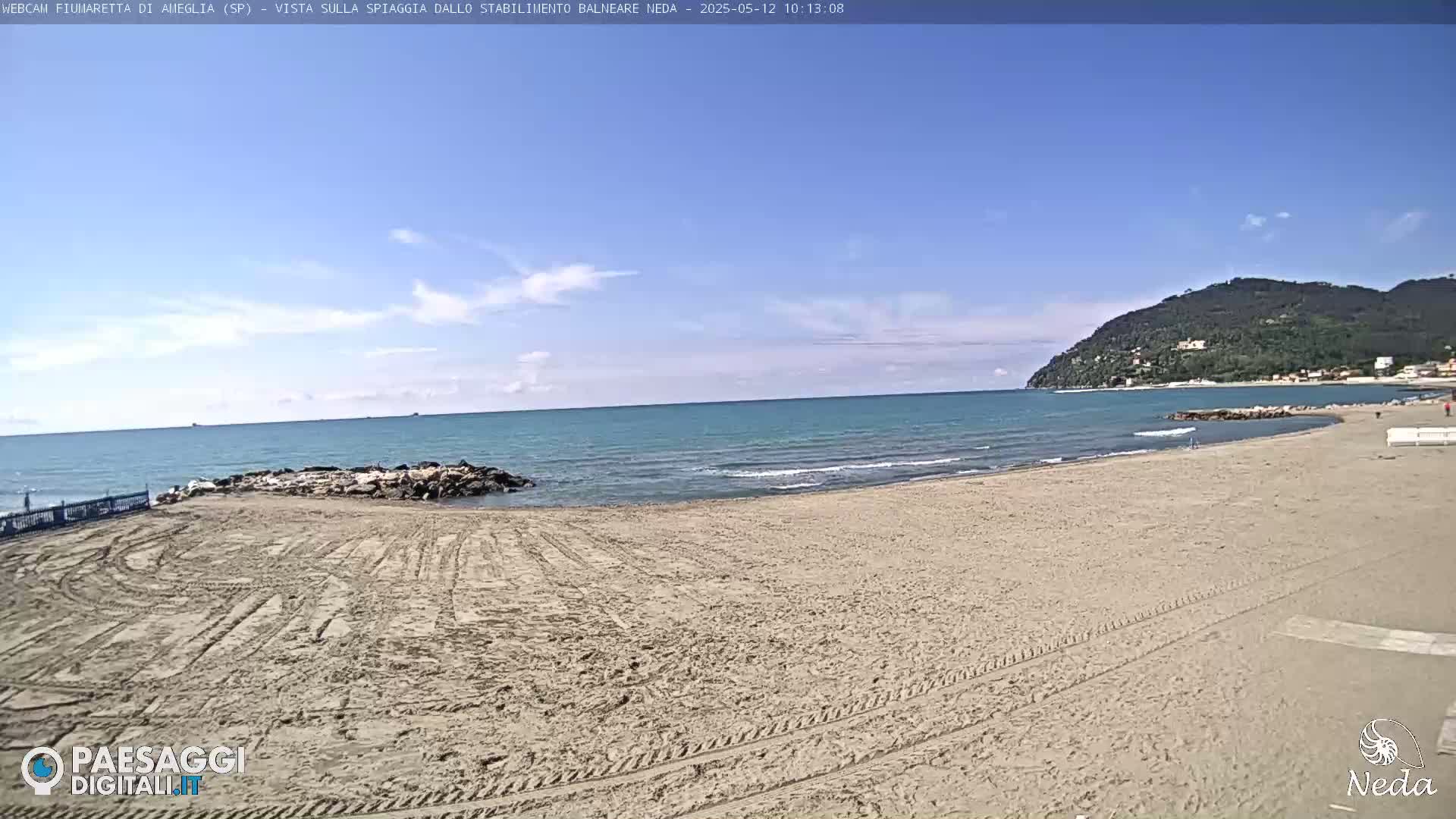 A sandy beach stretches towards a calm, teal ocean under a mostly clear, bright blue sky, with a distant hill visible in the background.