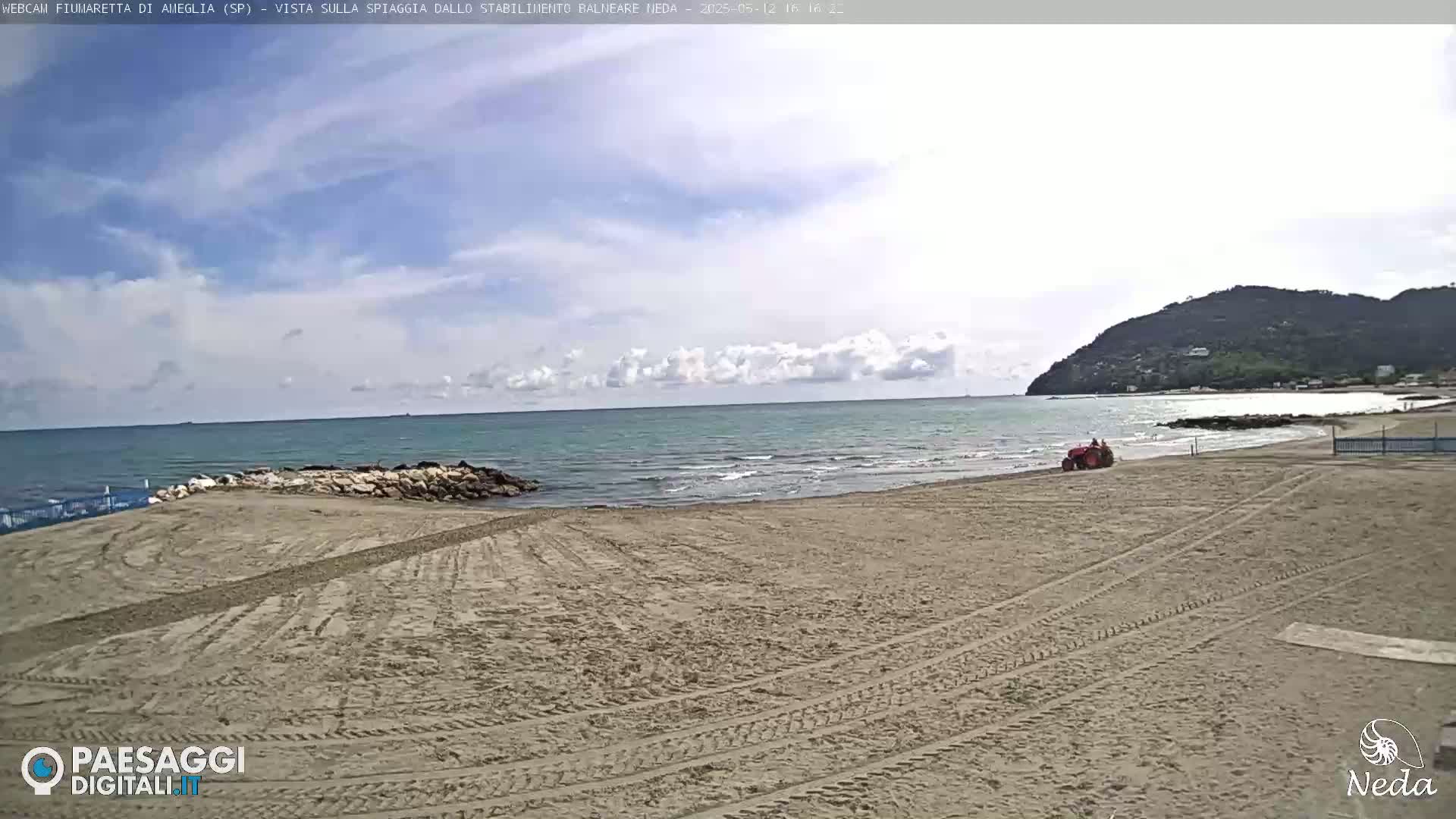 A sandy beach with a tractor on it, next to a calm sea under a partly cloudy sky, and a hill in the background.