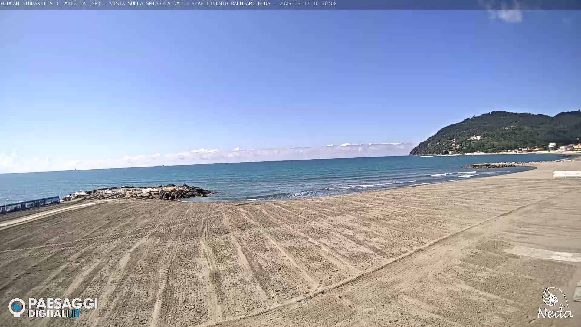 A sandy beach stretches toward a calm, blue ocean under a mostly clear sky, with a verdant hill visible in the distance.