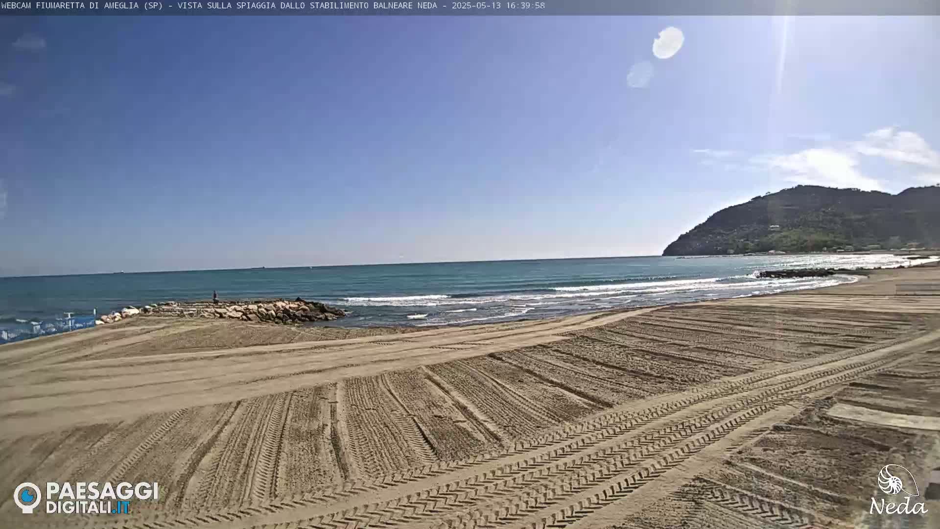 A sandy beach with tire tracks shows a calm ocean under a clear blue sky, and a hill is visible in the background.