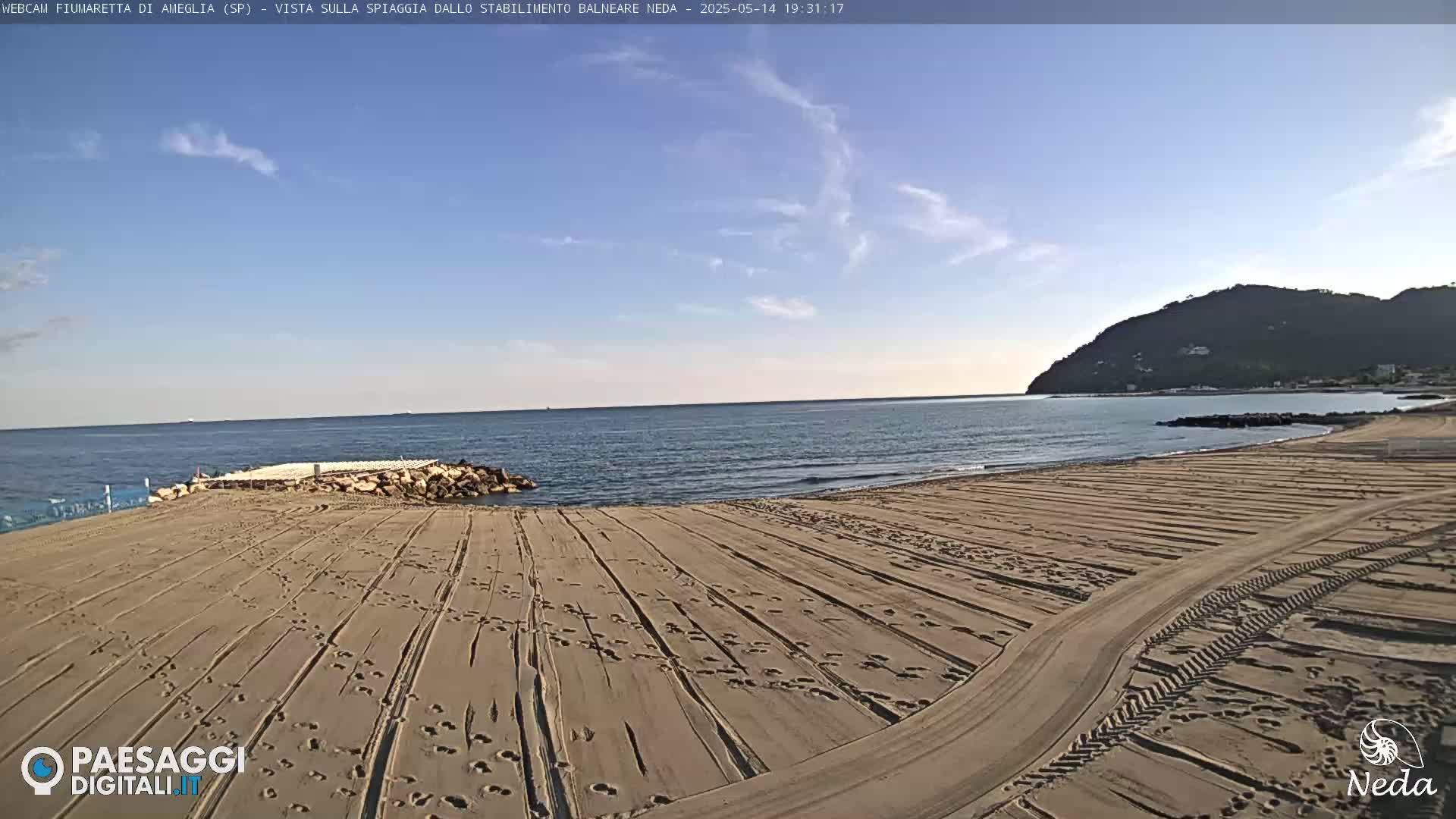 A sandy beach with tire tracks and footprints stretches toward a calm sea under a mostly clear sky, backed by a distant hill.