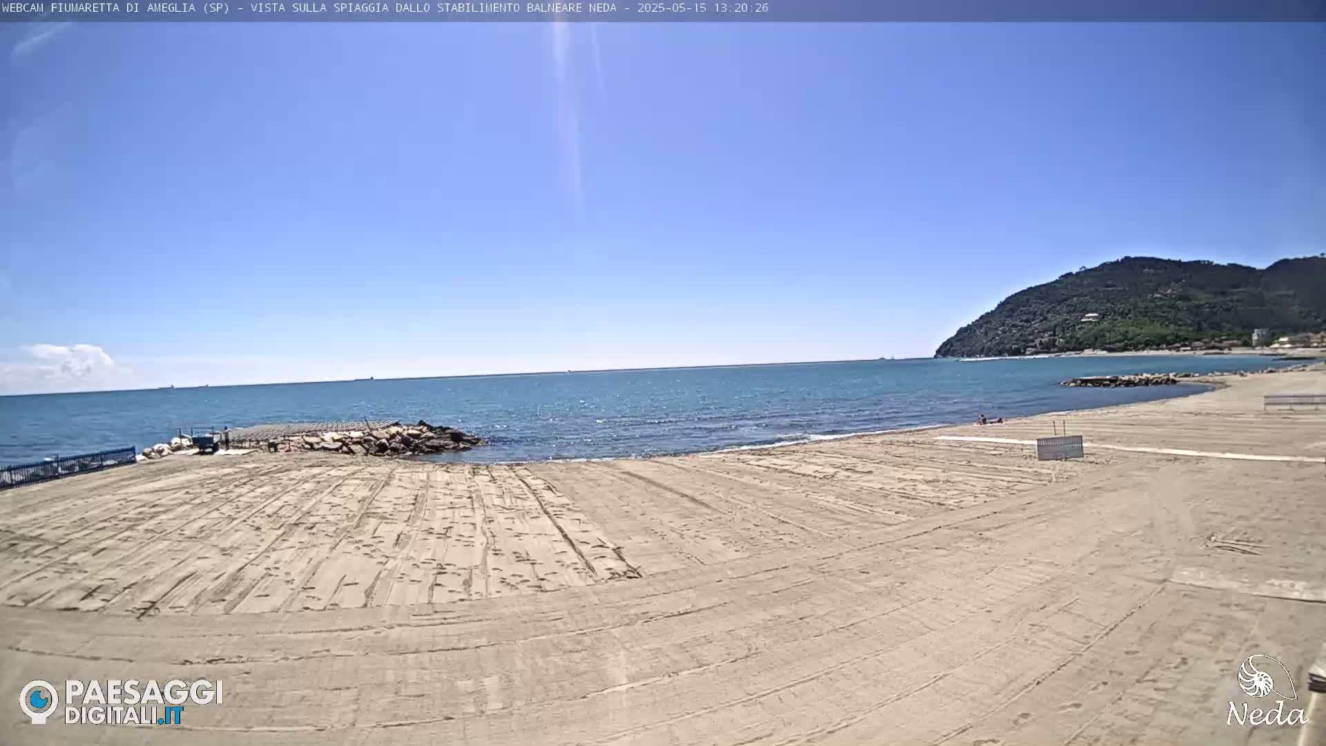 A mostly empty sandy beach stretches toward a calm blue sea under a clear blue sky, with a  green hill visible in the distance.