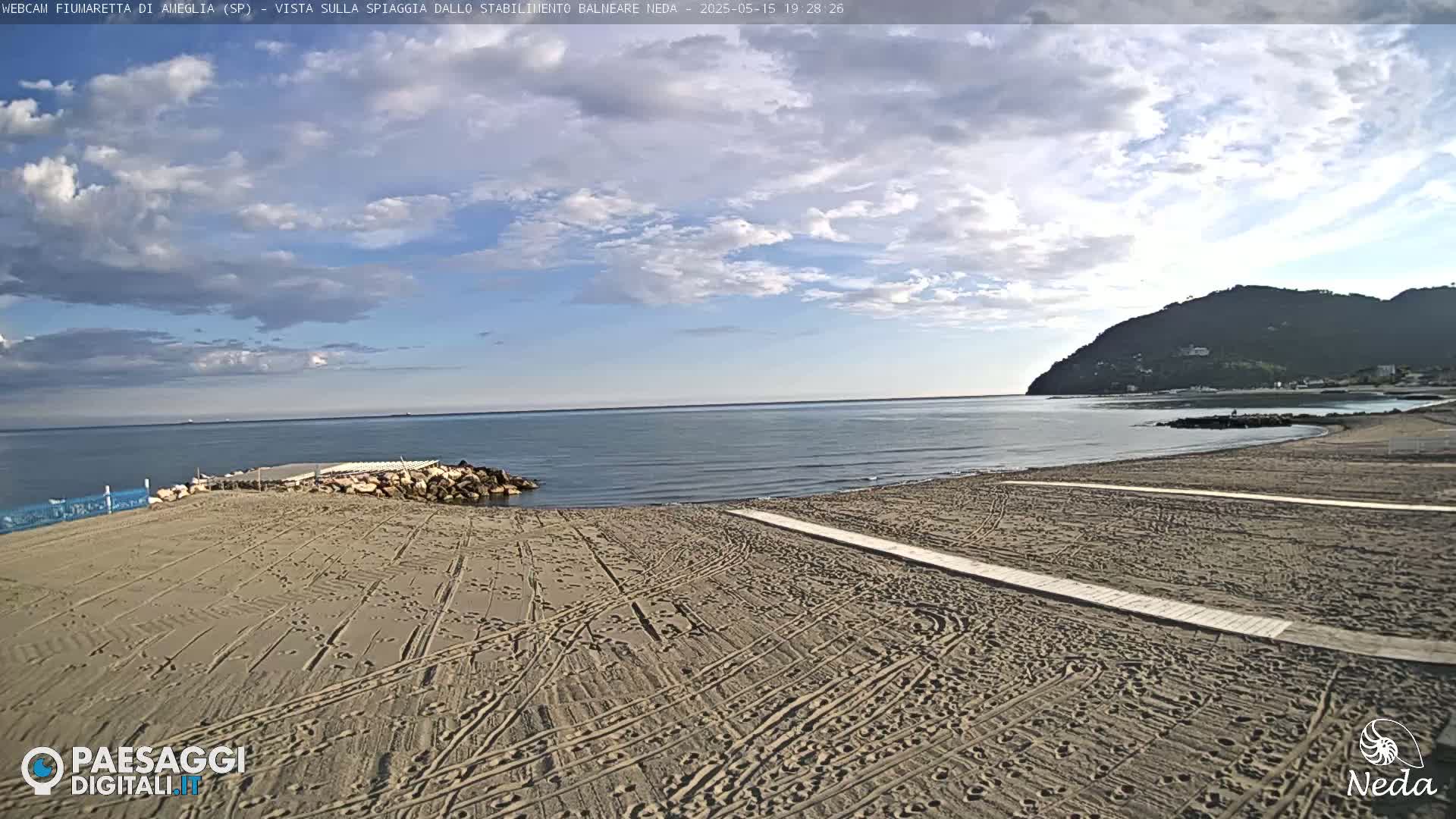 A sandy beach with tire tracks and walkways stretches towards a calm sea under a partly cloudy sky, backed by a distant hill.