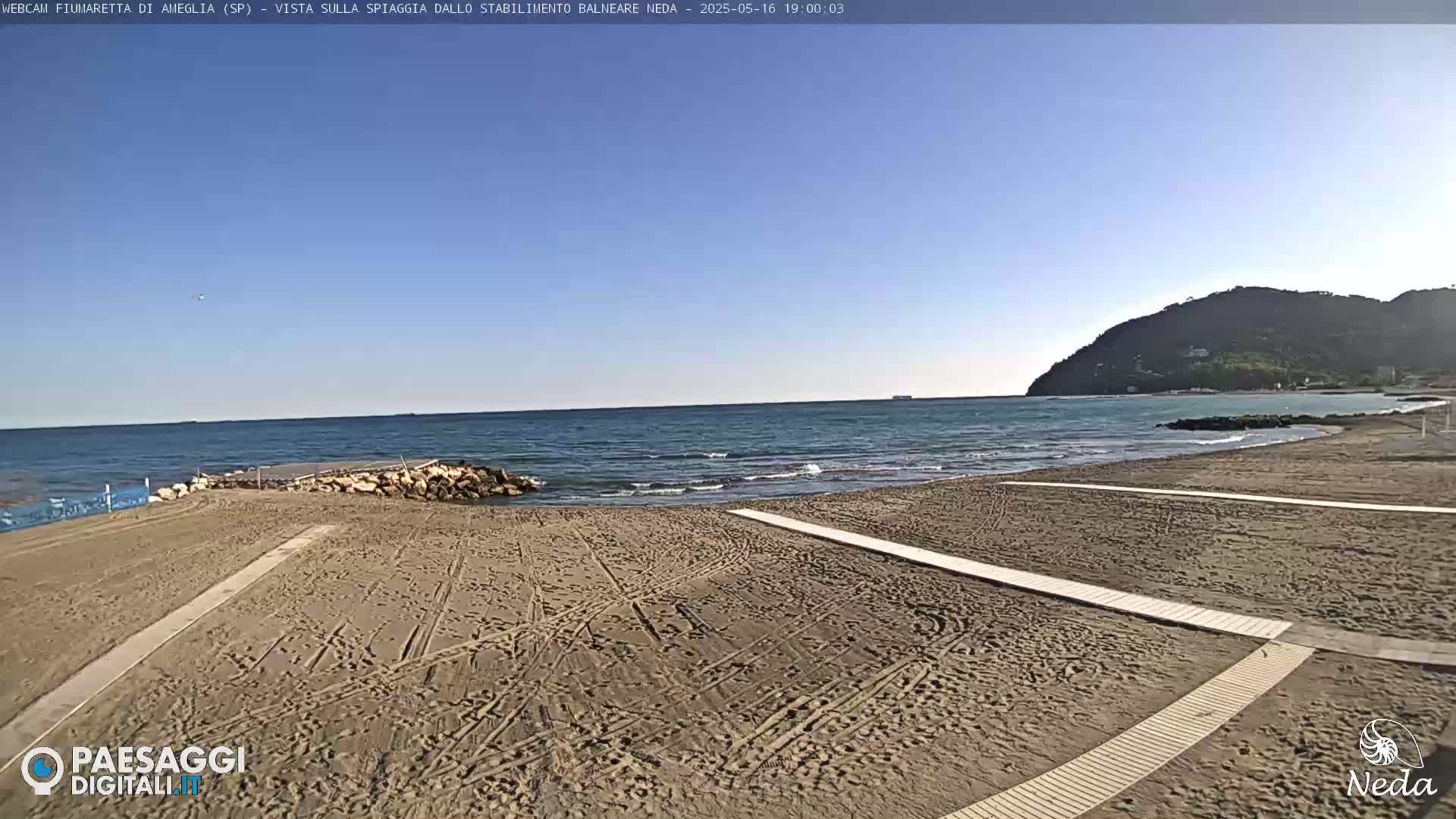 A sandy beach with wooden walkways stretches toward a calm sea under a clear blue sky, backed by a distant hill.