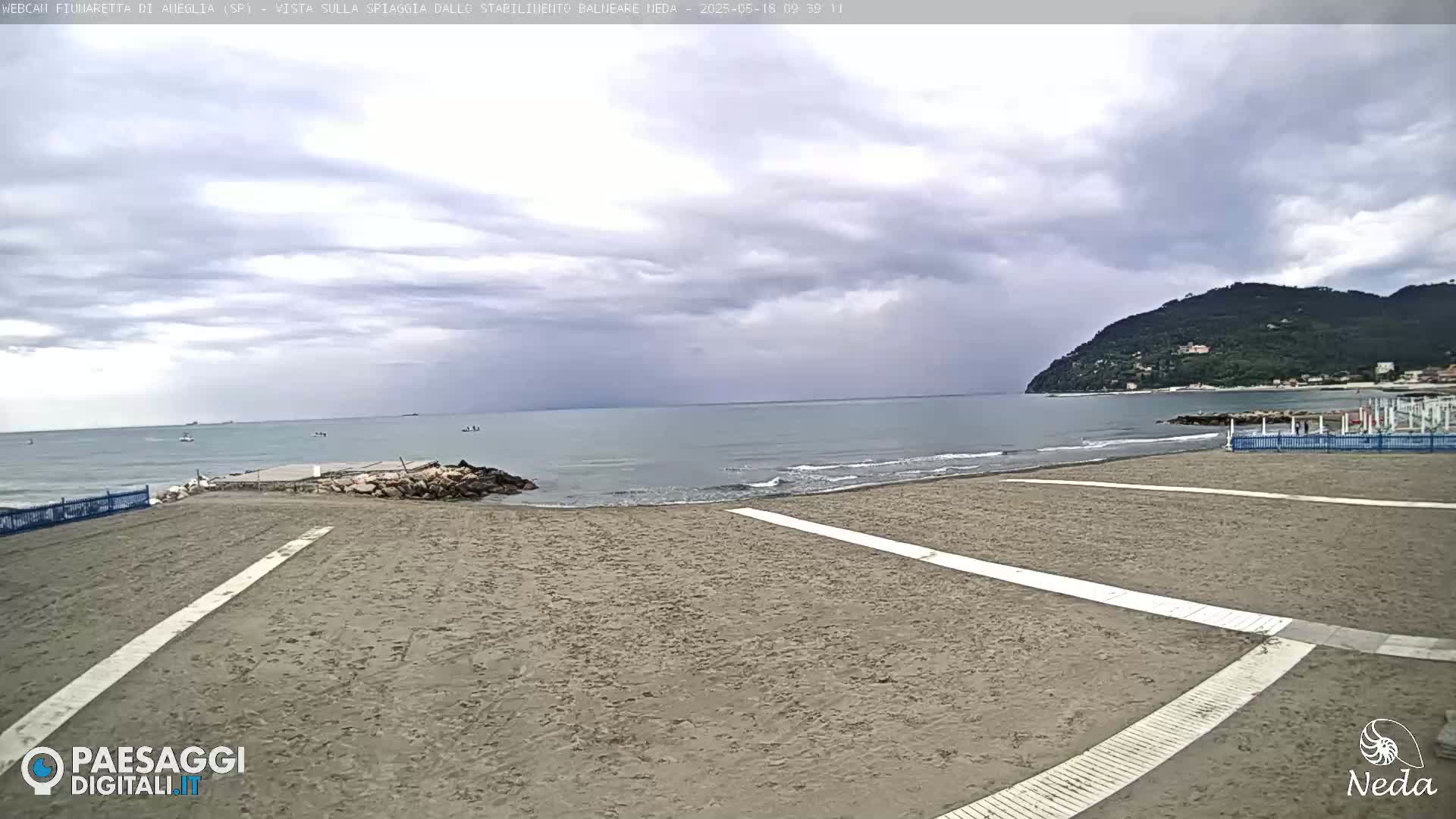 A sandy beach with a calm sea under a cloudy sky, backed by a hill covered in dark green vegetation.
