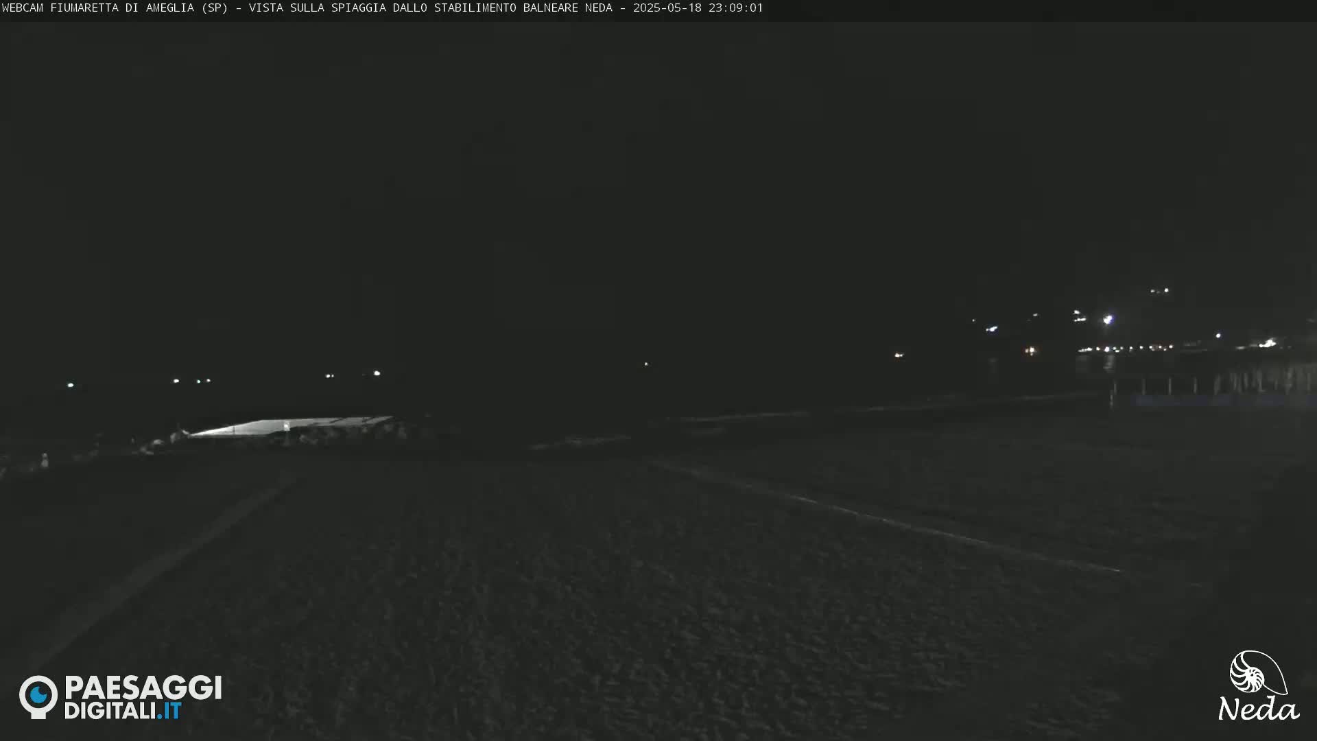 A dark, nighttime view of a beach with dimly lit structures in the distance.