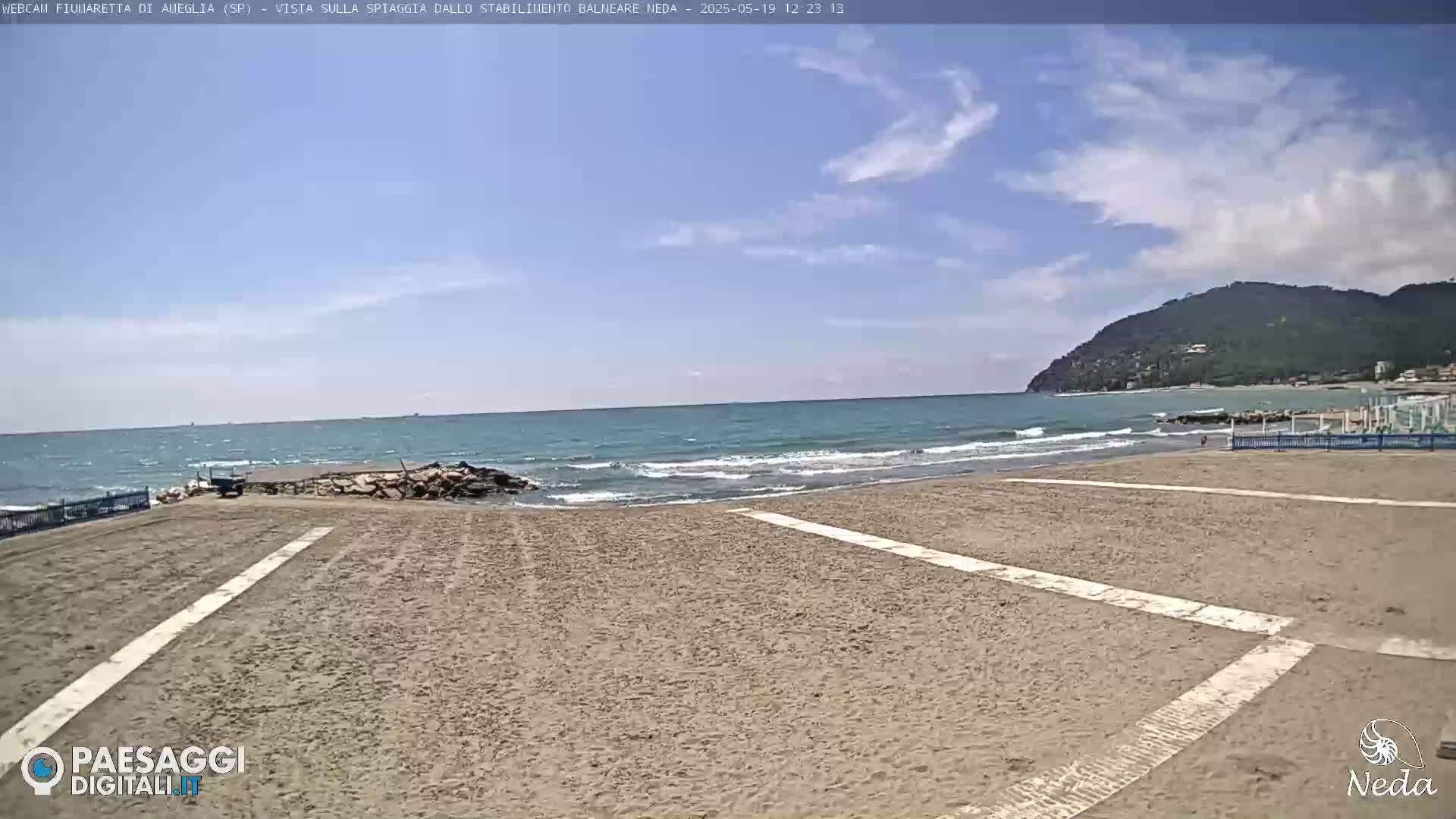 A sandy beach with a calm ocean under a mostly clear sky, backed by a distant mountain.