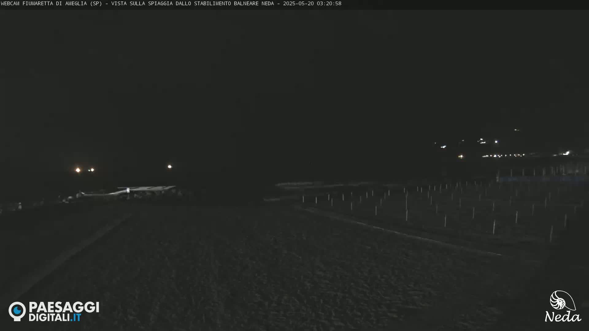A dark, nighttime view of a beach with sparsely lit structures in the distance.